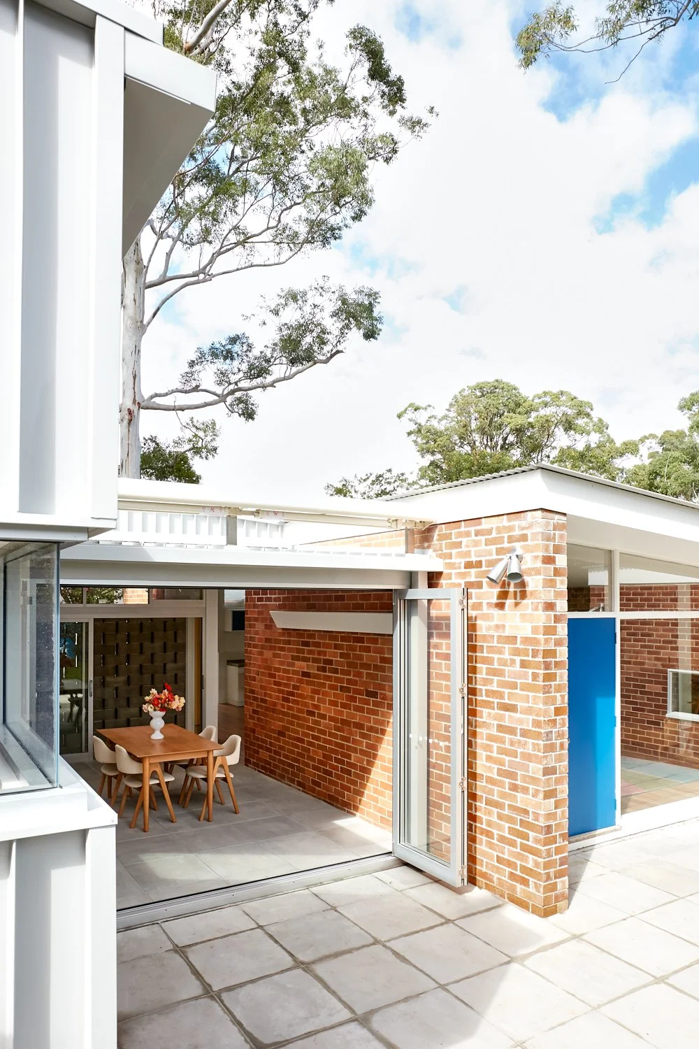 Outdoor patio area with brick walls, a wooden dining table with six chairs, a bouquet of flowers in a white vase, glass sliding door, and a clear sky with trees in the background.