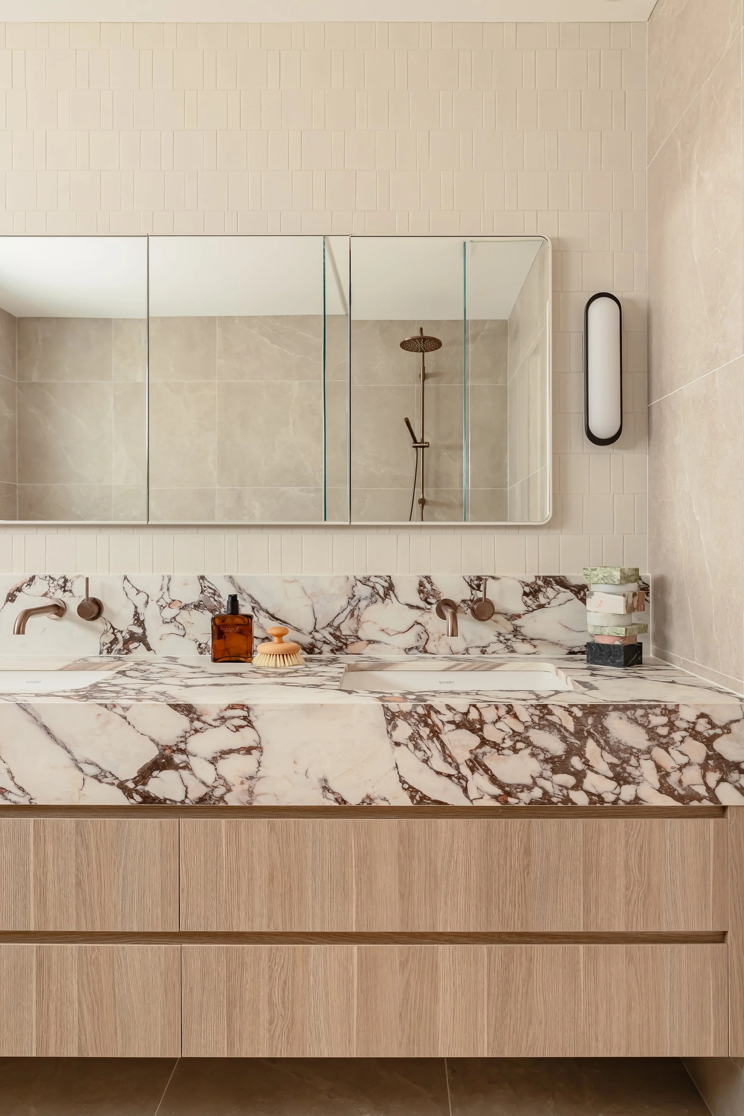 Modern bathroom with a marble double sink vanity, large mirror, wall-mounted faucets, and a walk-in shower with a rain showerhead, beige tiled walls, and a light fixture.