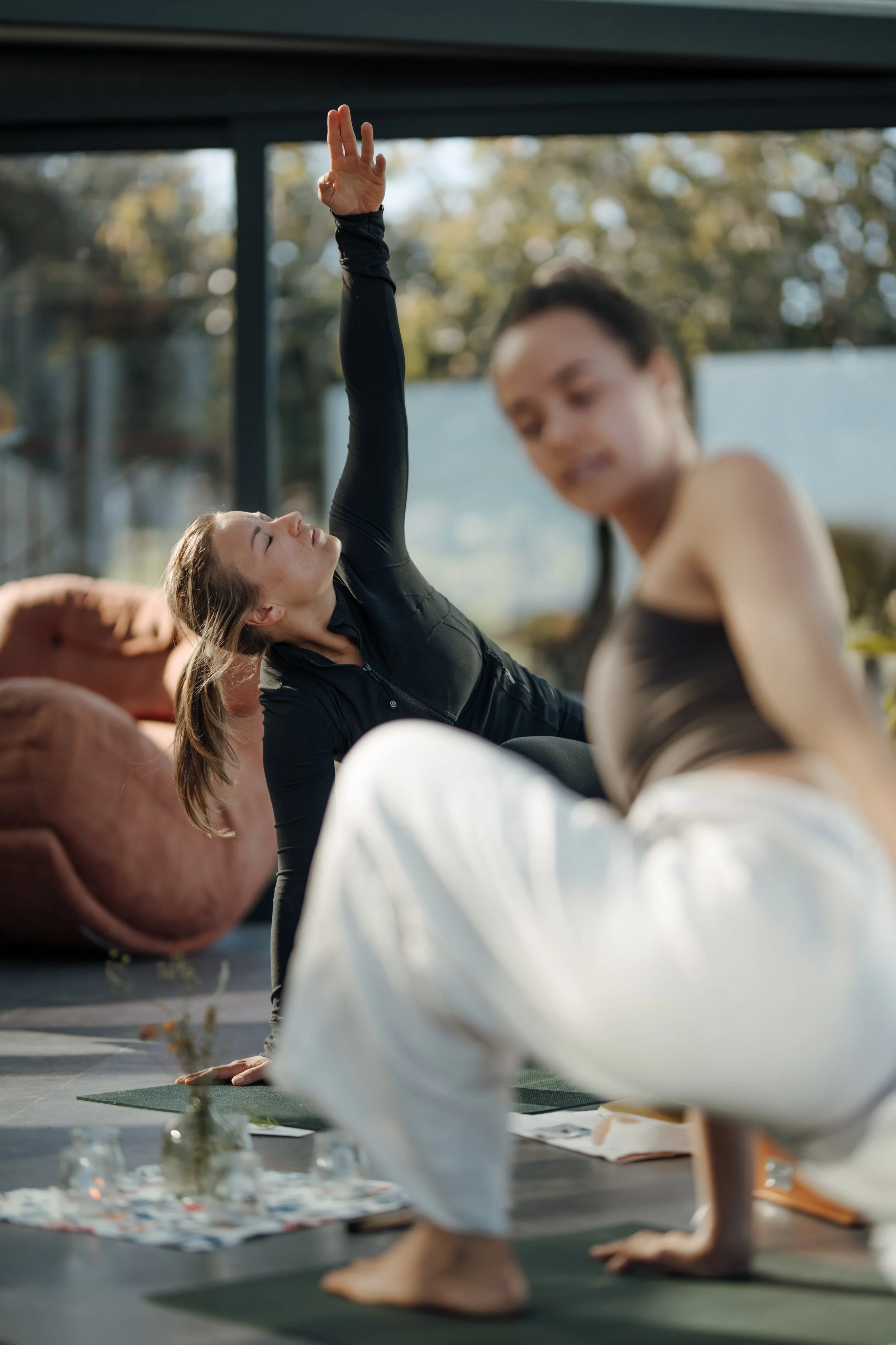 Two women practicing yoga indoors. One woman with dark hair and a black long sleeve top is extending her arm upward, and the other woman with darker hair is sitting cross-legged on a yoga mat in the foreground. The background shows large windows with a view of trees outside.
