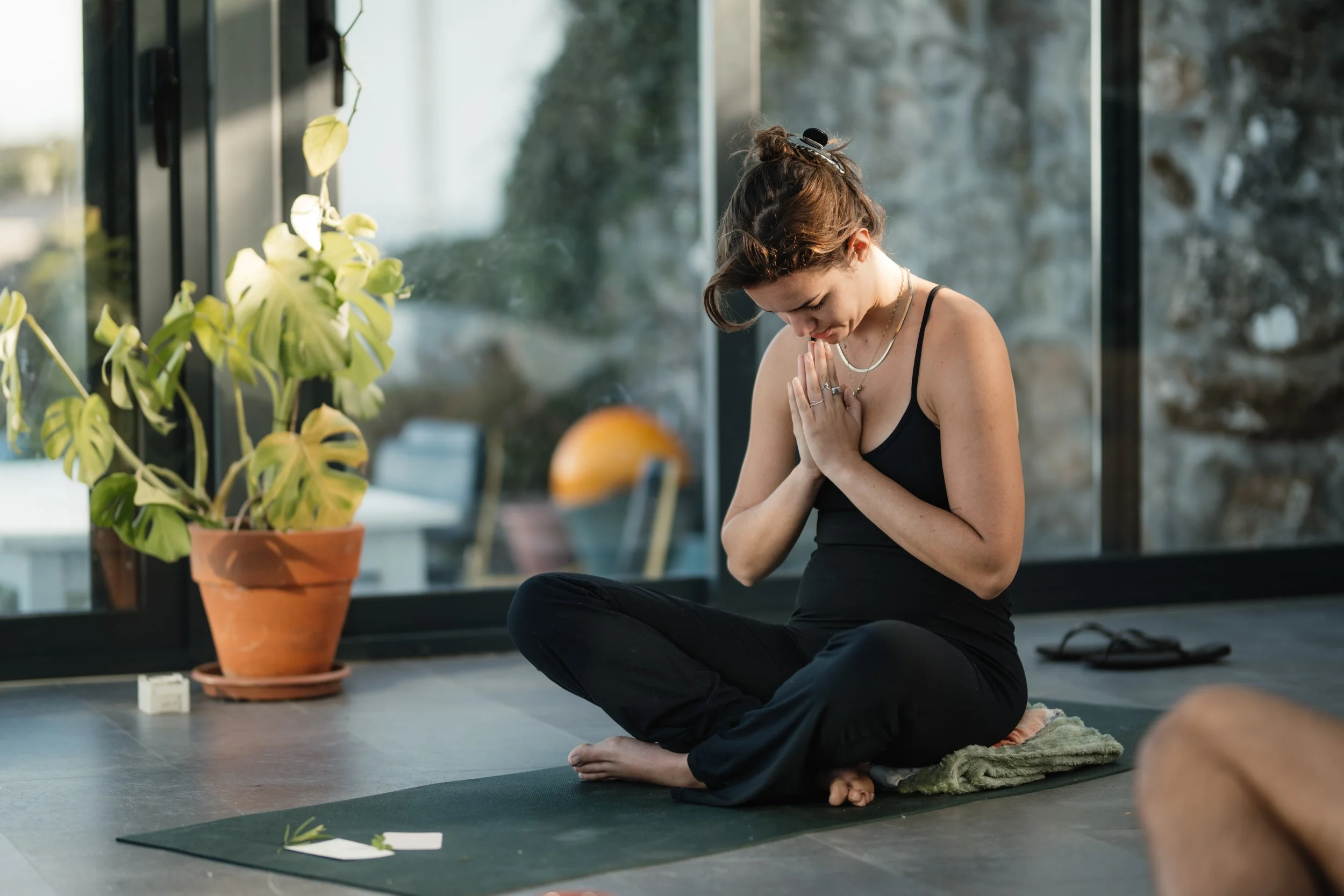 A woman practicing yoga or meditation indoors, sitting cross-legged on a mat, with her hands in prayer position and her head bowed. A large potted plant with green leaves is visible on her left.