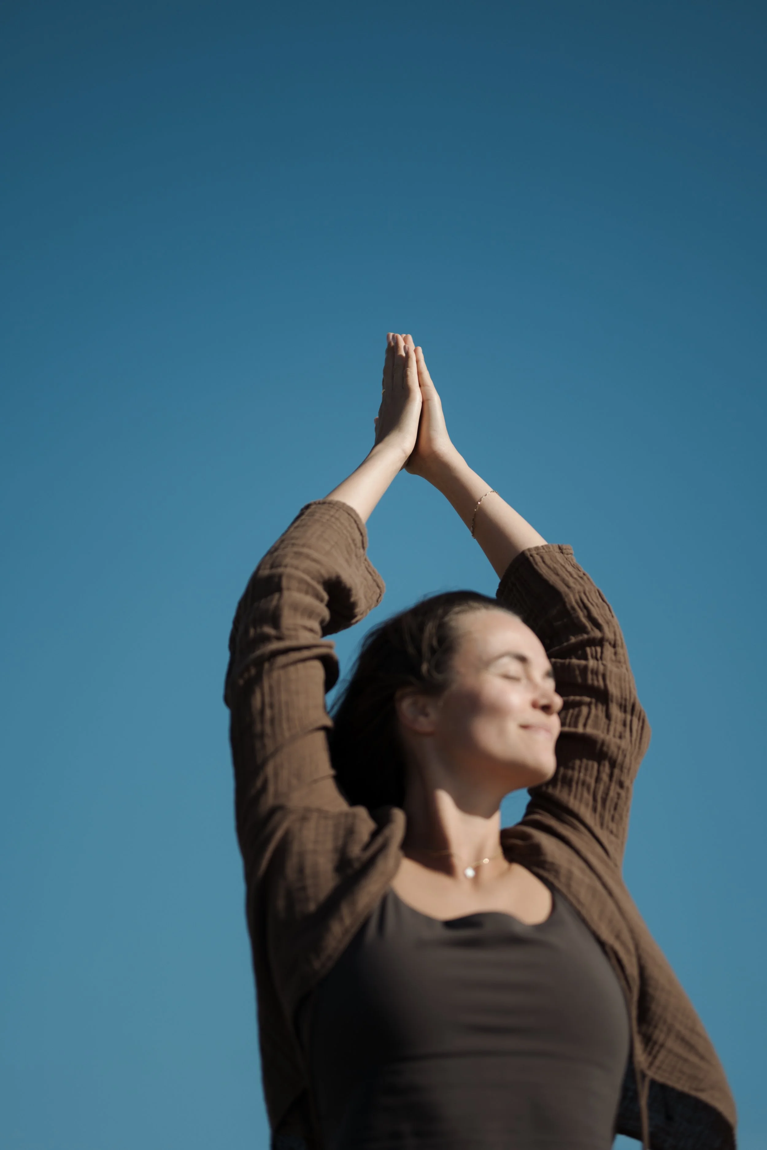 A woman standing outdoors with eyes closed, smiling, palms pressed together above her head, against a clear blue sky.