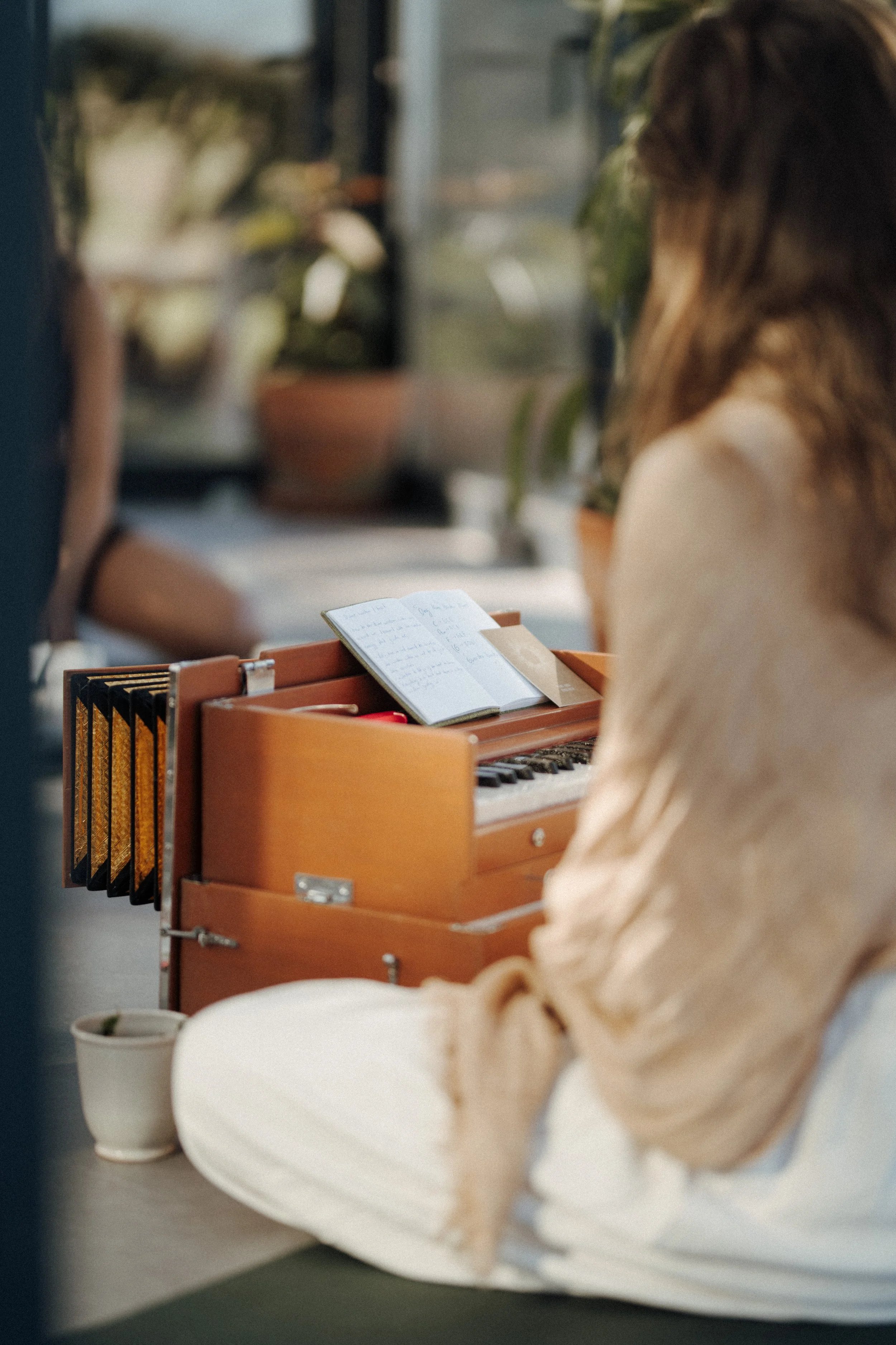 A woman is sitting at a table in a cafe, playing a small wooden piano. She has long hair and is wearing a beige blazer. There is an open notebook with handwritten notes on the piano. In the background, there are potted plants and blurred details of the cafe interior.