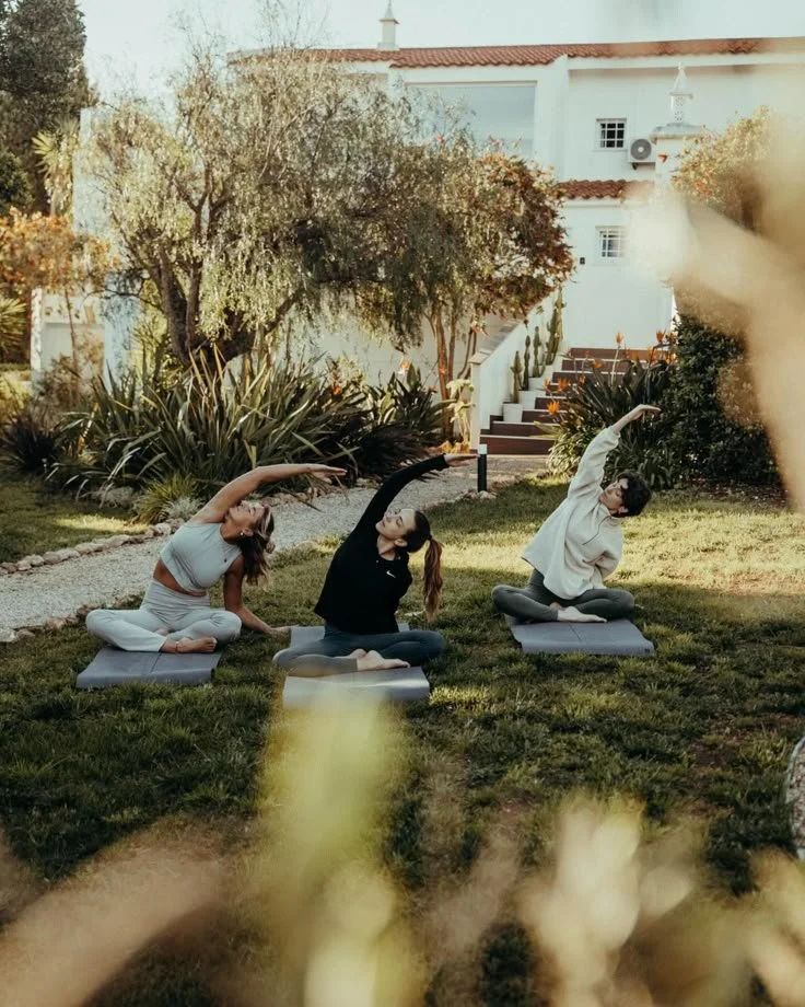 Three women practicing yoga outdoors on mats in a garden with trees and a white building in the background.