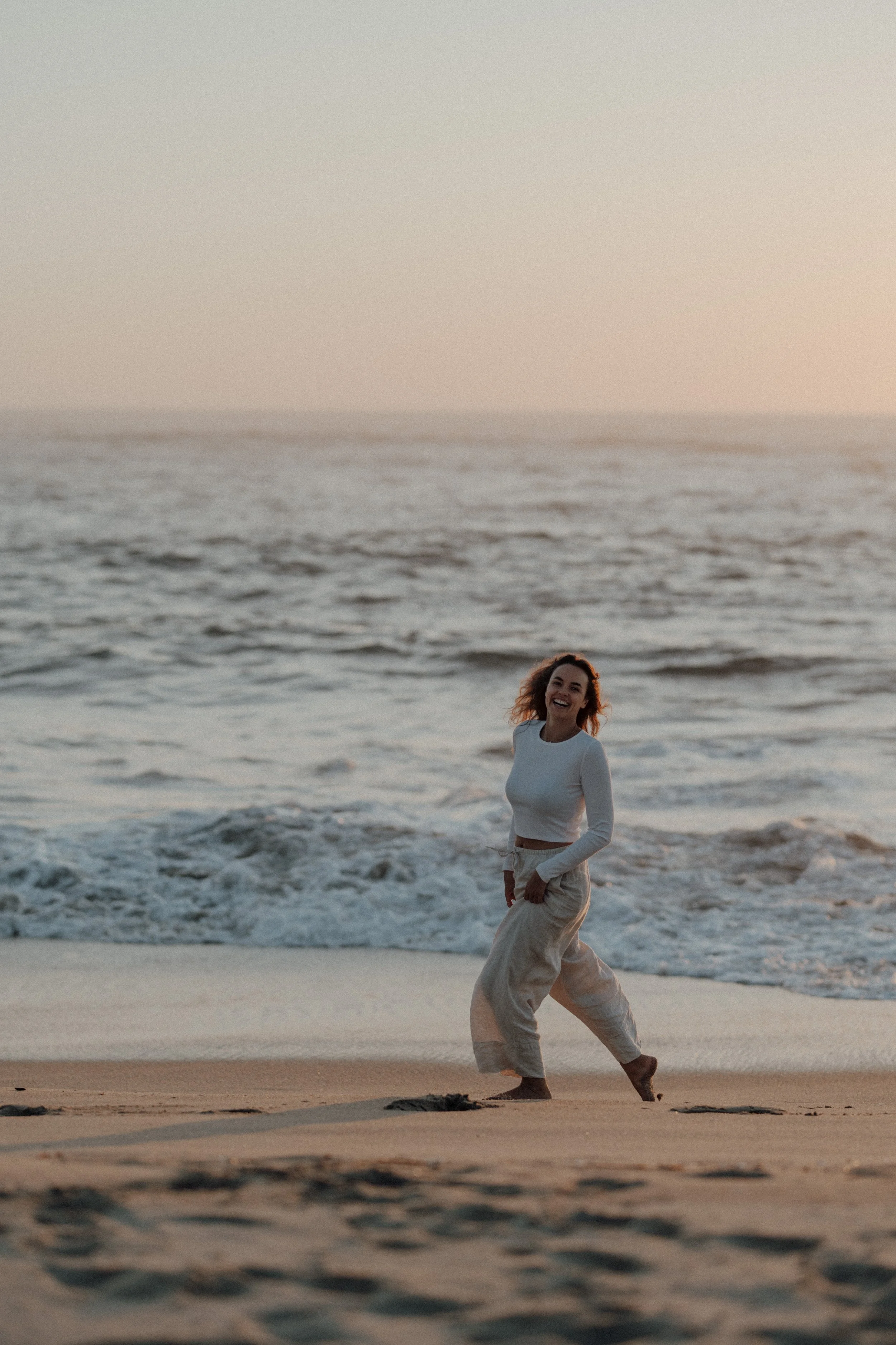 A woman smiling and walking along the beach at sunset, with the ocean waves in the background.