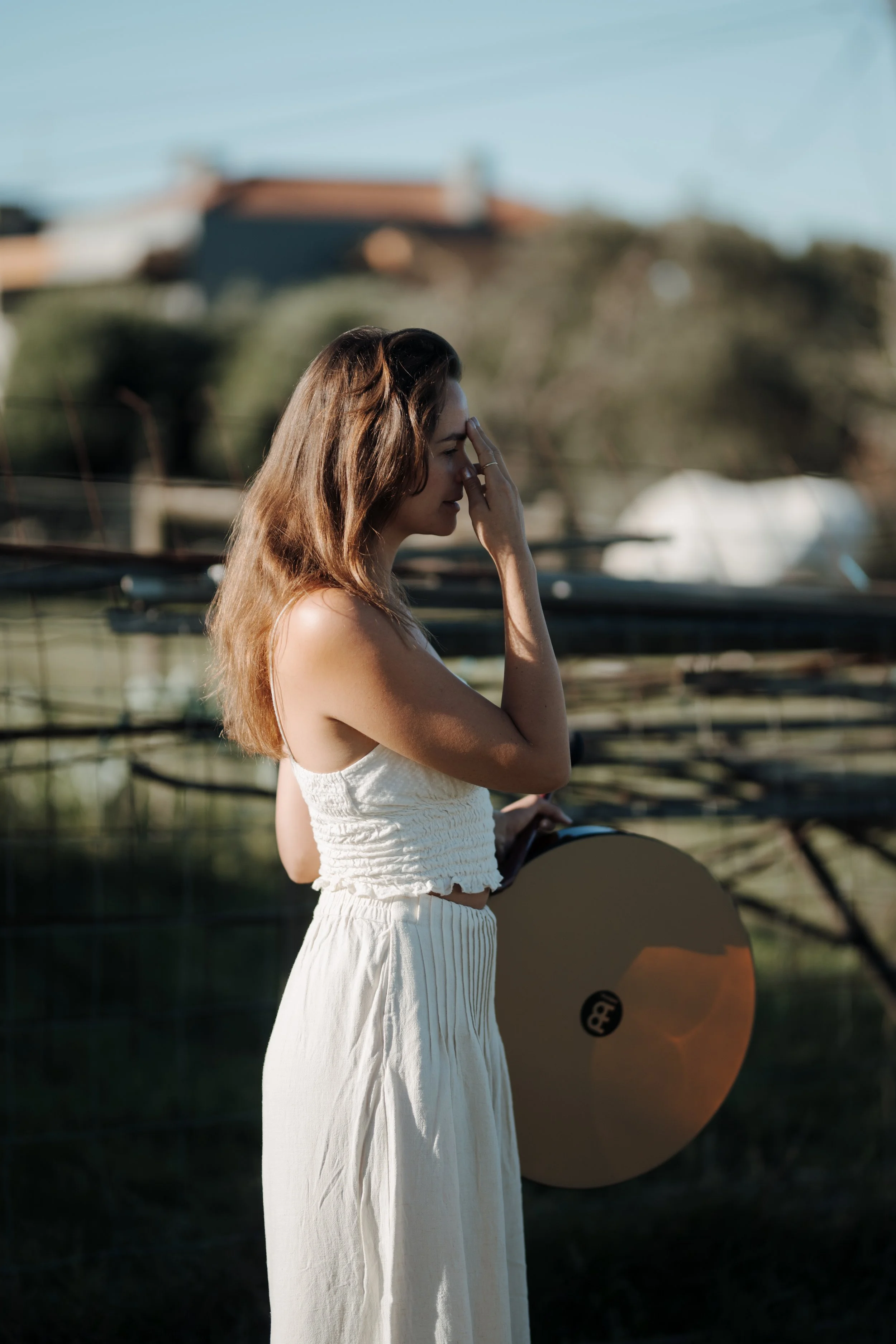 A woman with brown hair wearing a white sleeveless top and white skirt, holding a skateboard, standing outdoors near a boat, with a blurry background of trees and houses.