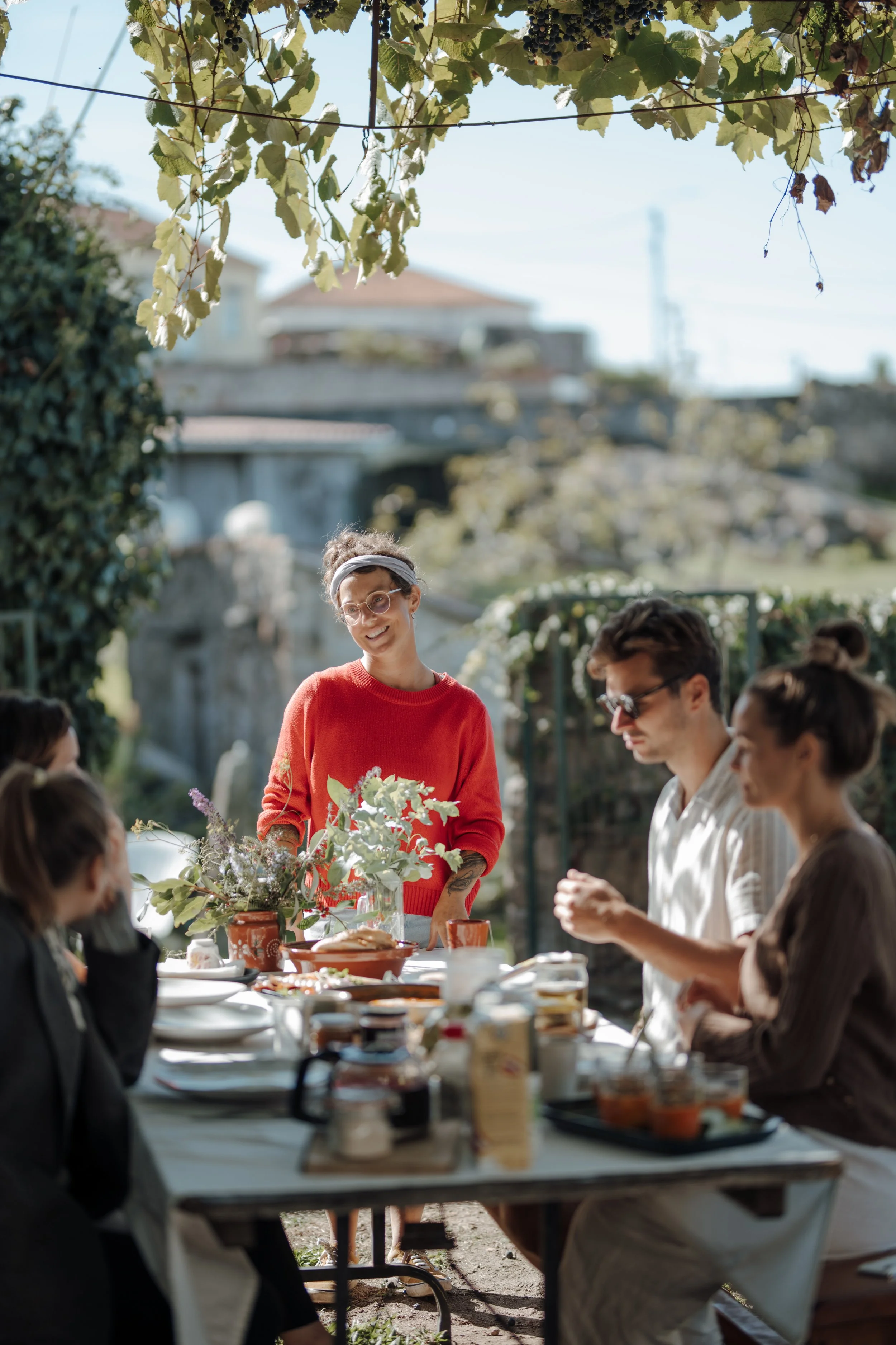 People gathered around a table outdoors during daytime, with a woman in a red sweater standing and smiling at the group, surrounded by laid-out food and a natural setting with trees and buildings in the background.