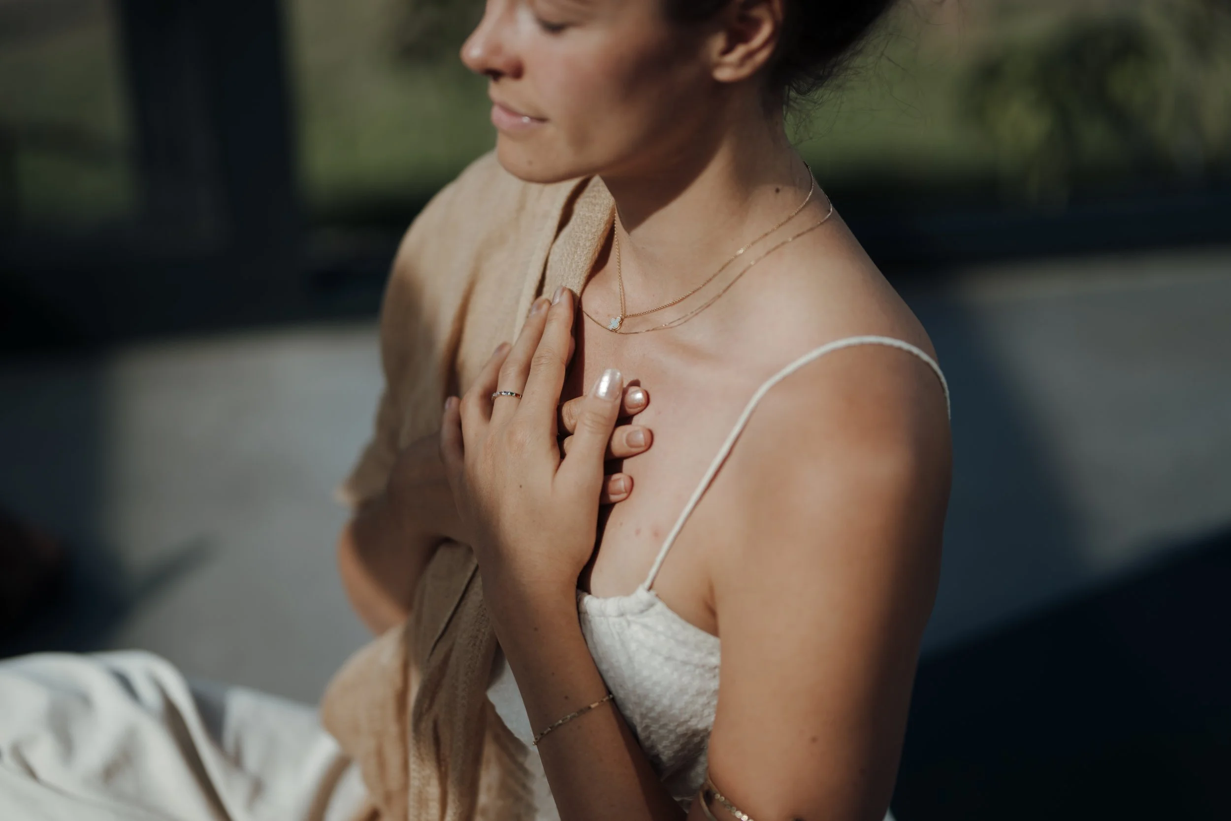 A woman with her eyes closed, touching her chest with her hand, wearing layered necklaces and a light-colored dress, sitting outdoors with greenery in the background.