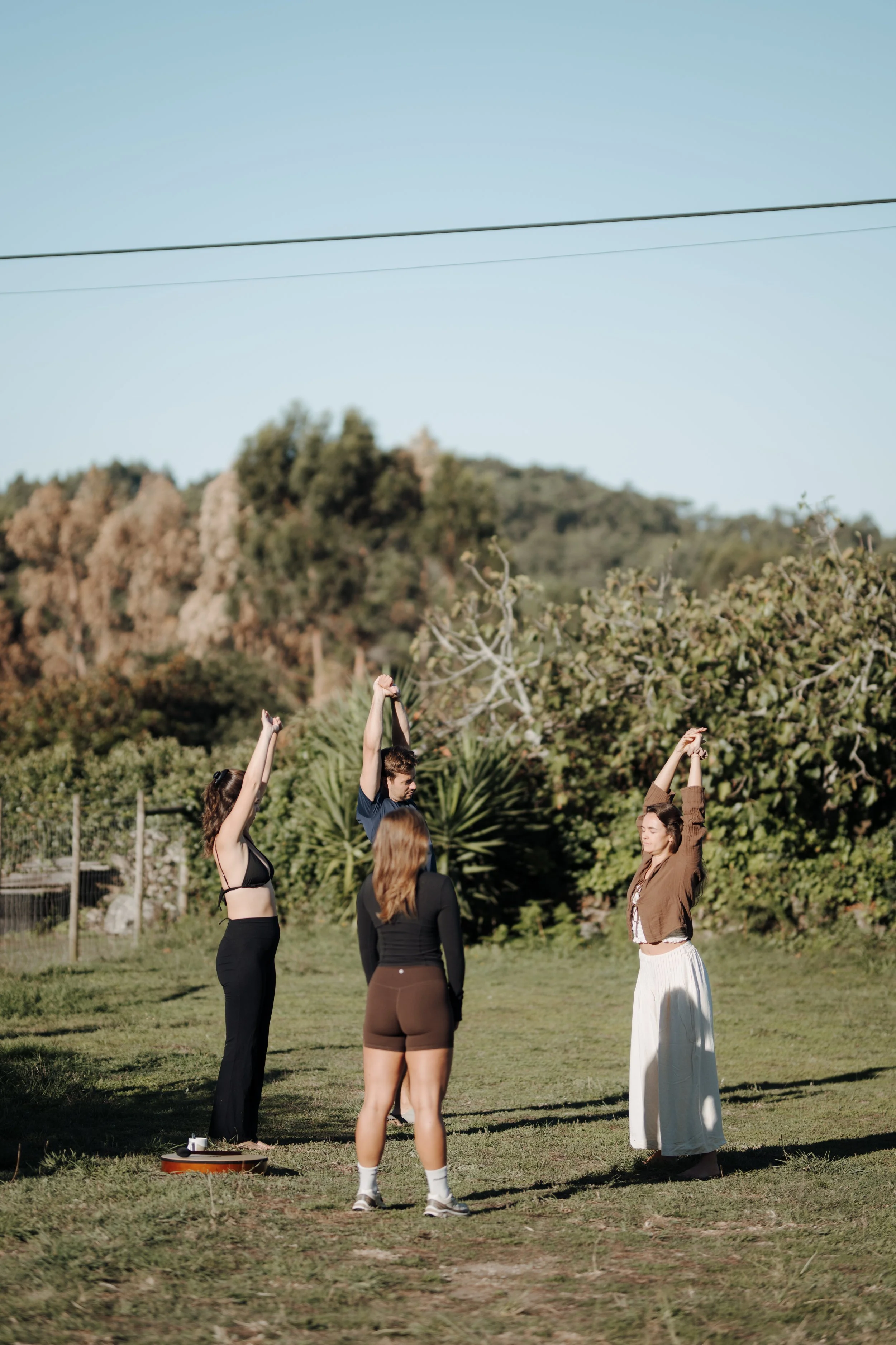 Four women standing in a circle outdoors with trees and bushes in the background, engaging in a yoga or stretching activity, with one woman raising her arms above her head.