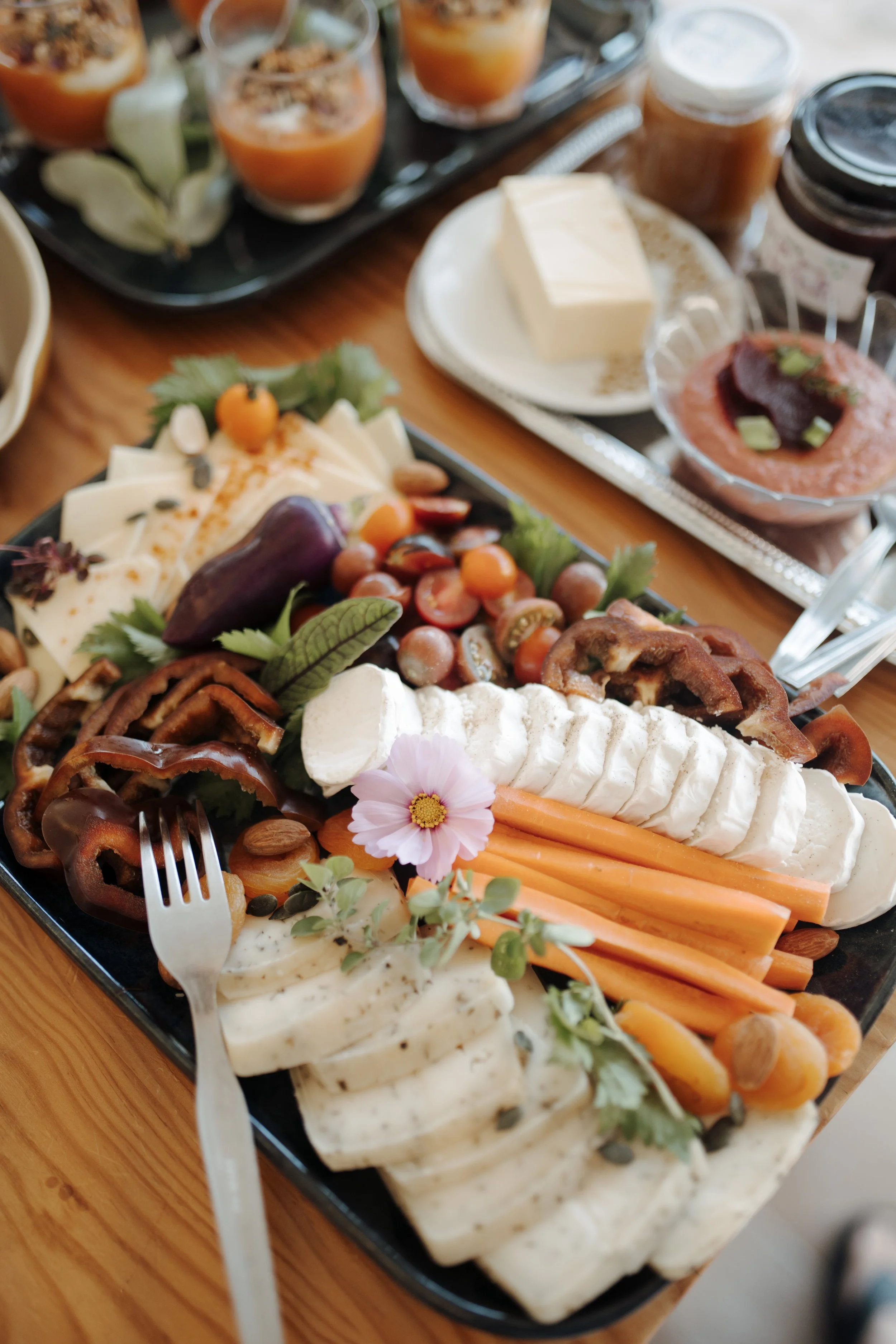 A cheese and vegetable platter with carrots, cherry tomatoes, cheese slices, and herbs, served with a fork and garnished with flowers, topped with various dips and condiments in small jars and bowls.