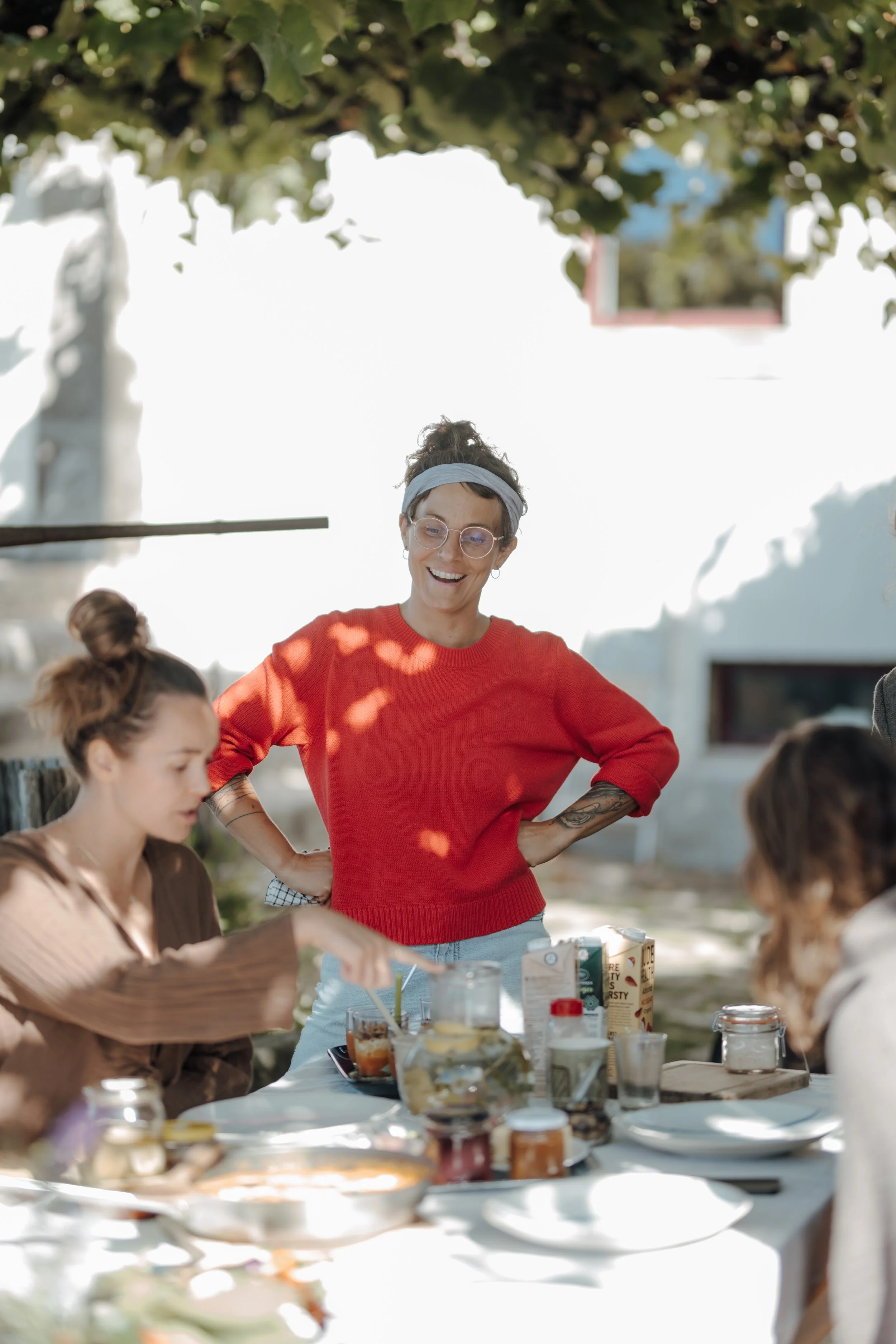 A woman with short hair, glasses, and a blue headband smiling and standing with hands on hips outdoors during daytime behind a table with food and drinks. Several people, mostly women, are seated at the table.