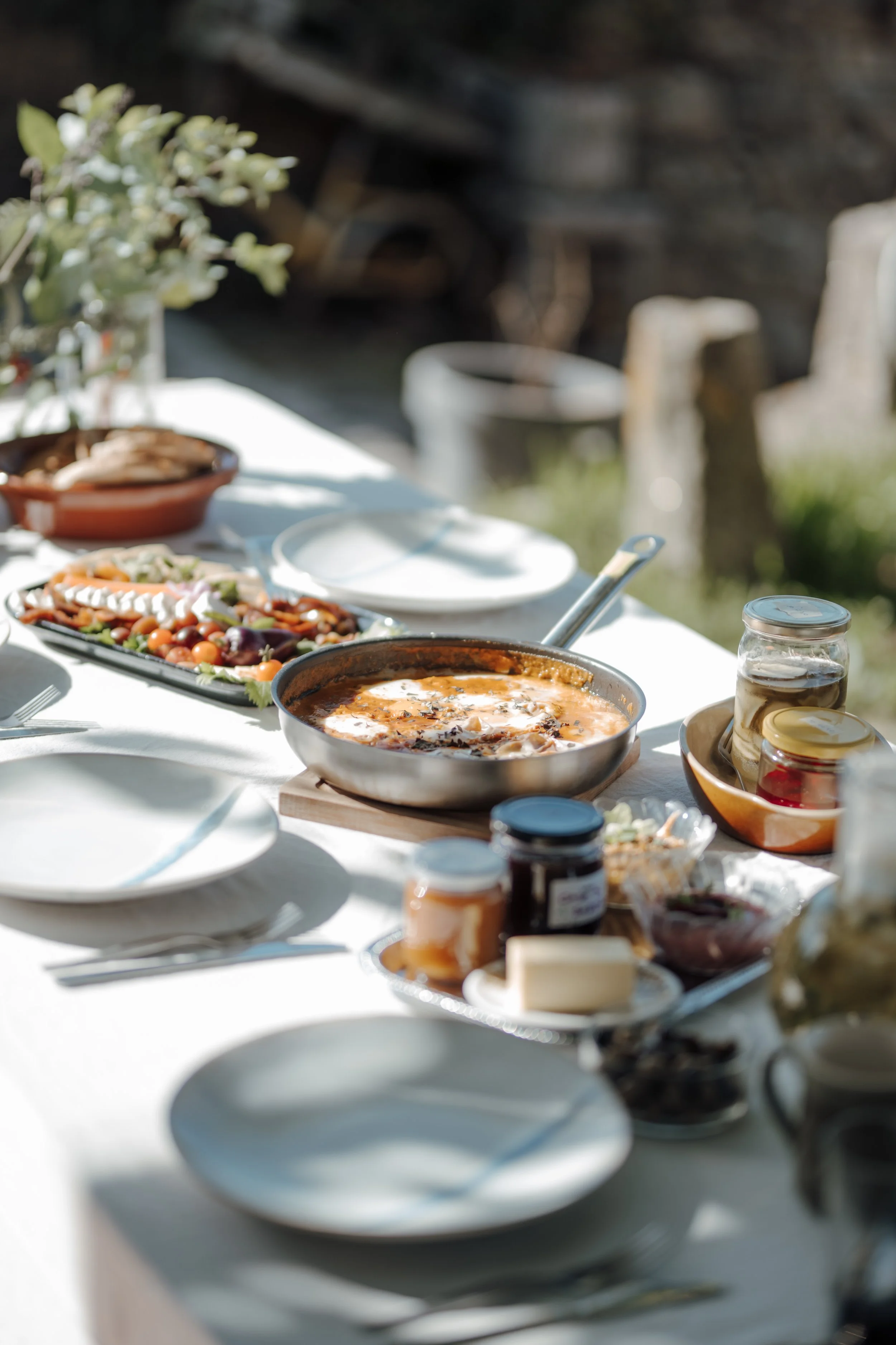 A table set outdoors with various dishes including a mixed salad, a bowl of soup, and condiments, with blurred background and sunlight.