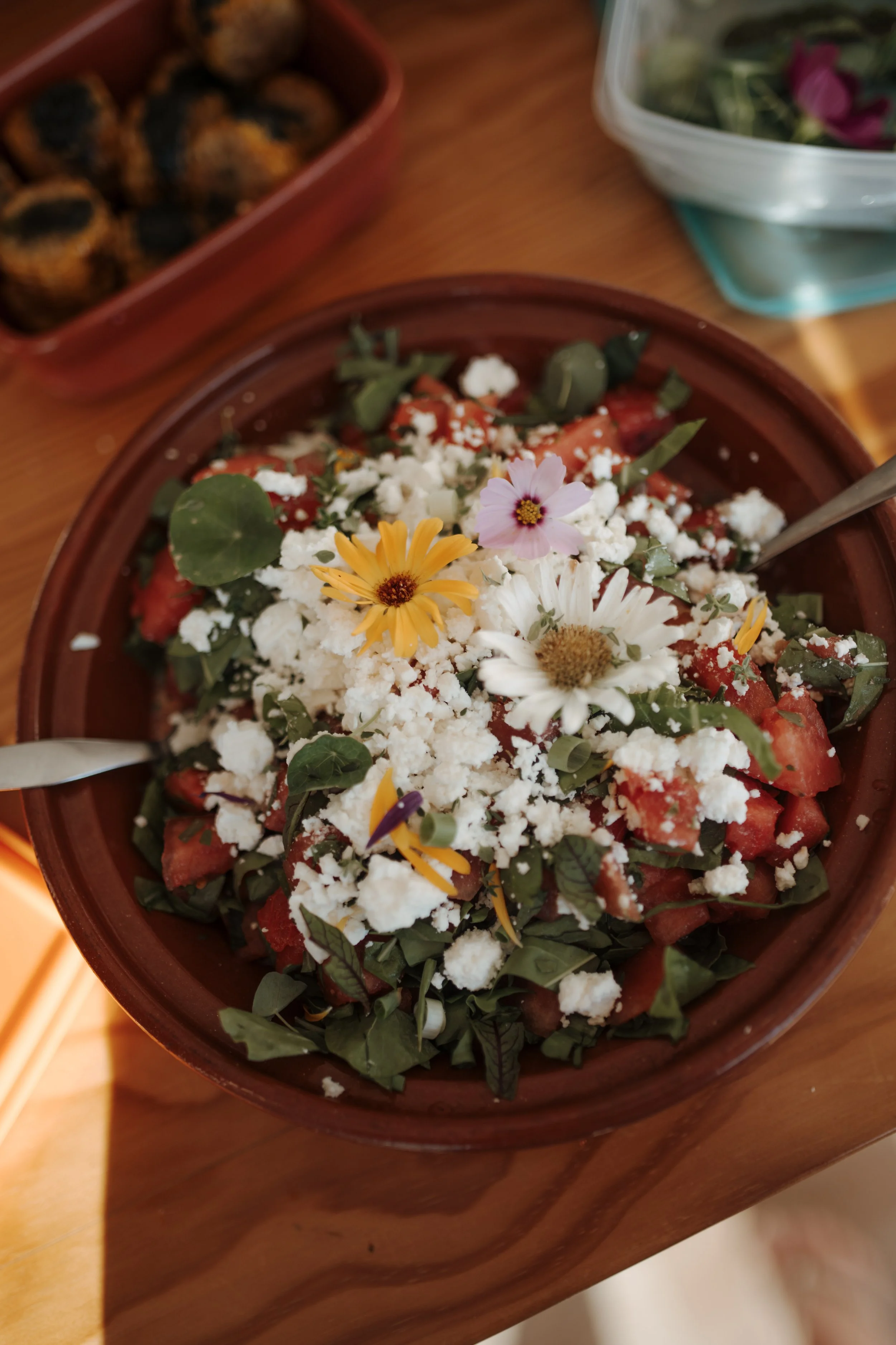 A bowl of chopped watermelon topped with crumbled white cheese and edible flowers, next to a container of salad and a tray of roasted or grilled food on a wooden table.