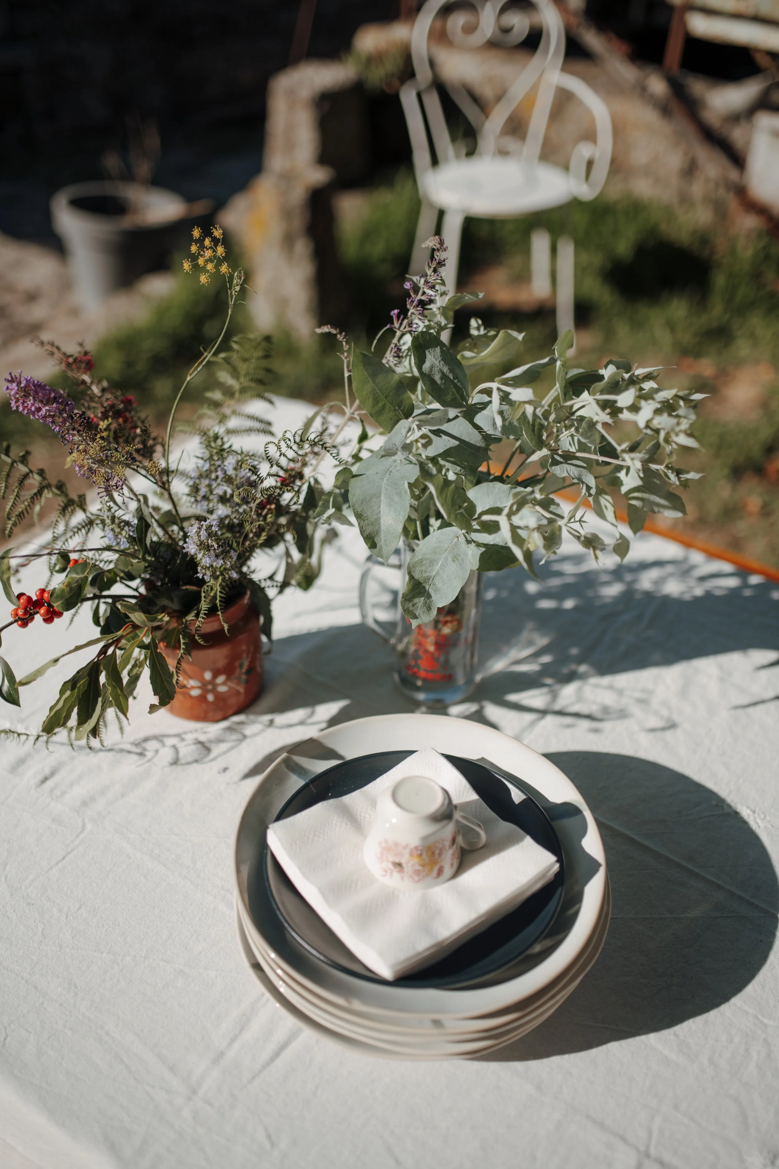 A table with a floral centerpiece in a glass vase, a stack of plates with a napkin and a teacup on top, and a potted plant in the background outdoors on a sunny day.