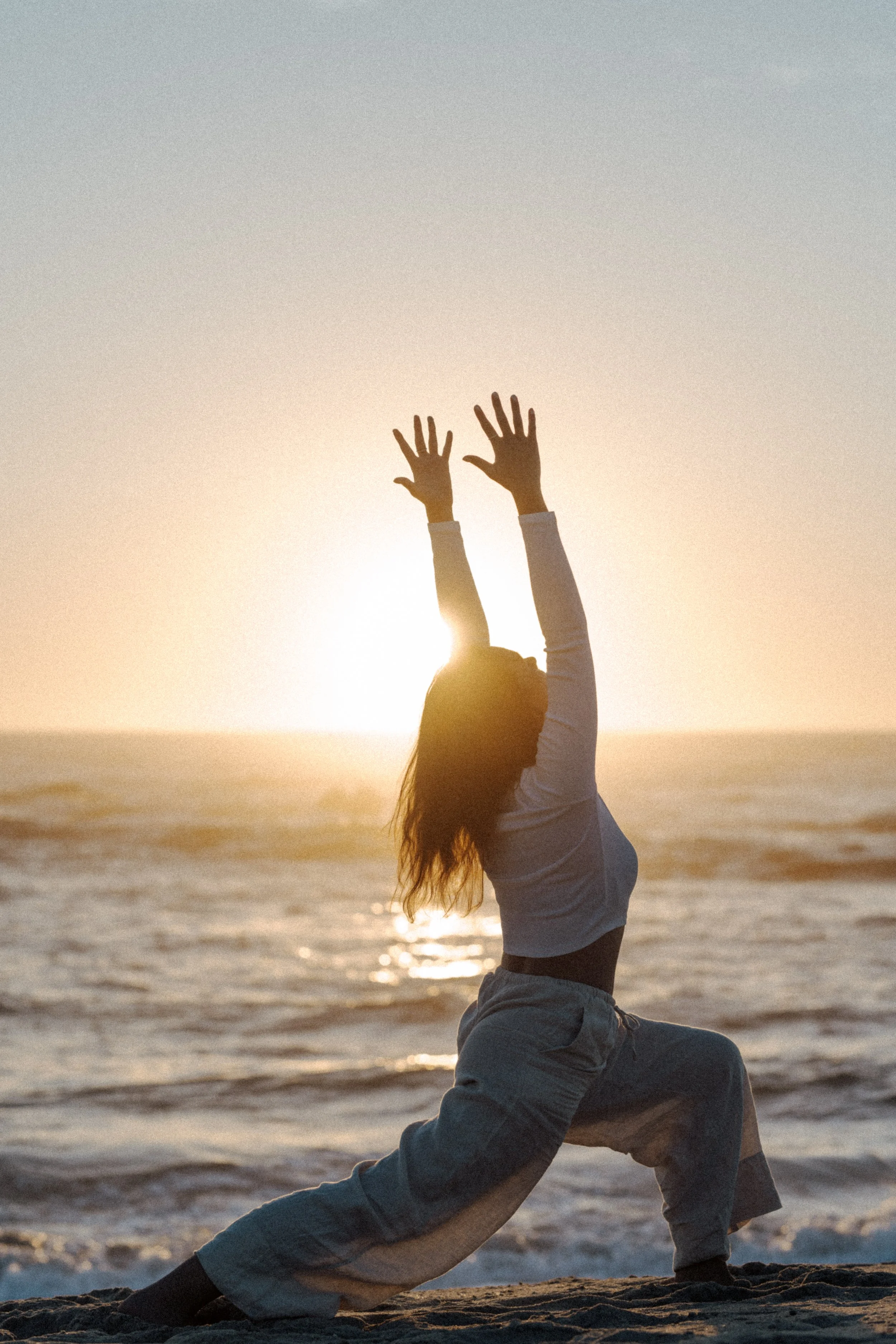 A person performing a yoga pose on the beach during sunset, balancing on one hand with the other hands raised towards the sky.