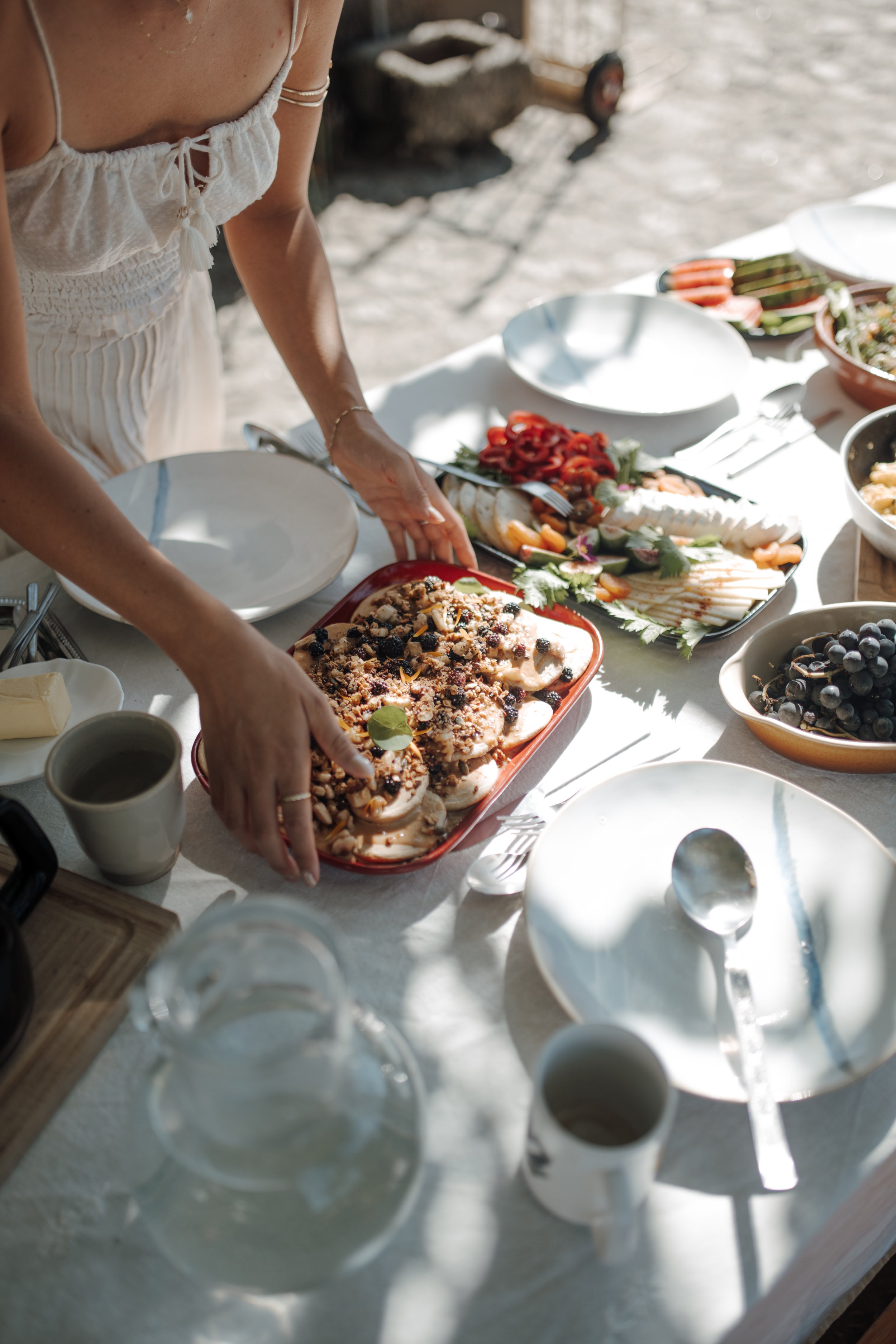 Person in a white dress standing at a table filled with various dishes, including a tray with baked goods, bowls of grapes, and plates of sliced vegetables, outdoors in a sunny setting with sand and shadows.