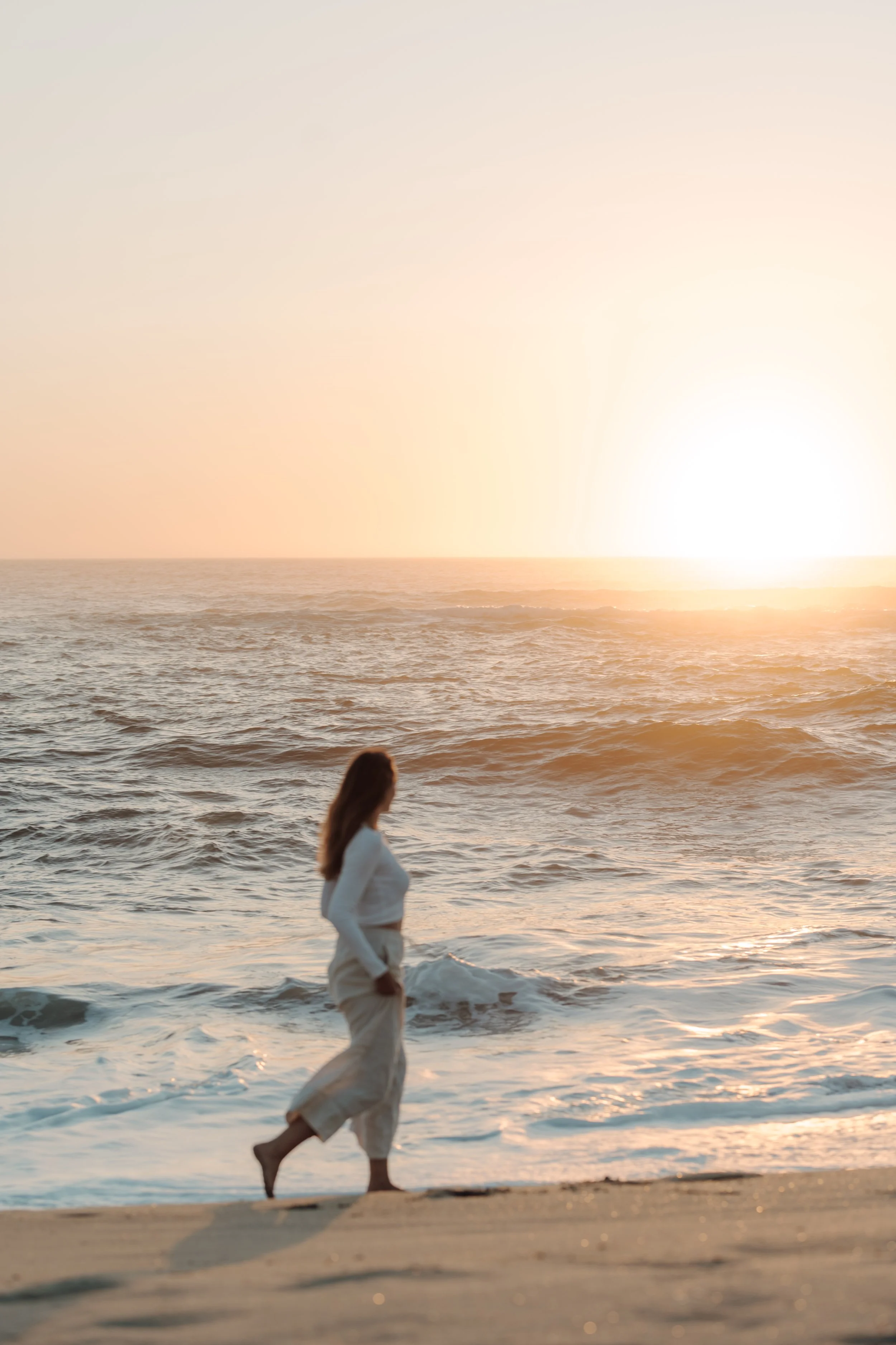 A woman in white clothing walking along the beach at sunset with the ocean in the background.