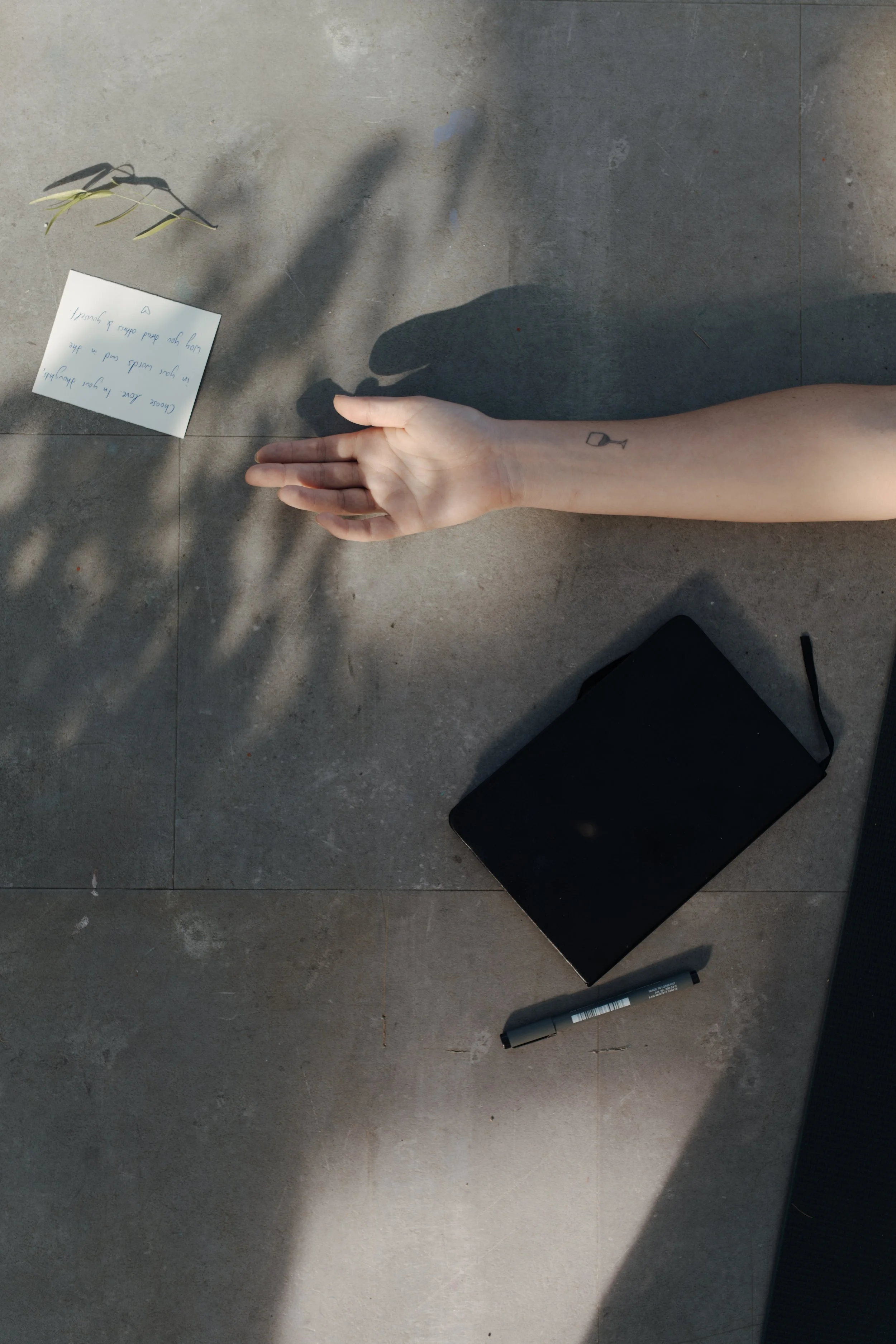 A person's arm with a small tattoo of a wine glass, laying on a tiled floor, with a pen, a black notebook, a white note card with handwritten text, and a small sprig of leaves nearby. The person's shadow is cast on the floor.