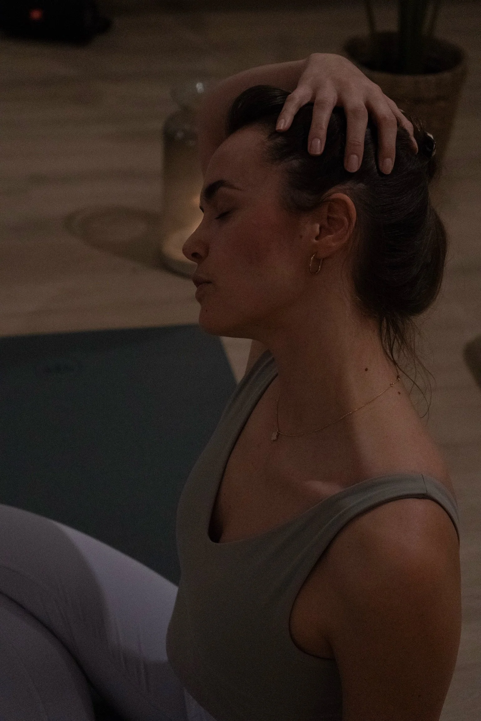 A woman with dark hair tied back, wearing a gray tank top, standing in front of a washing machine, with her eyes closed and one hand on her head in a moment of reflection inside a home.