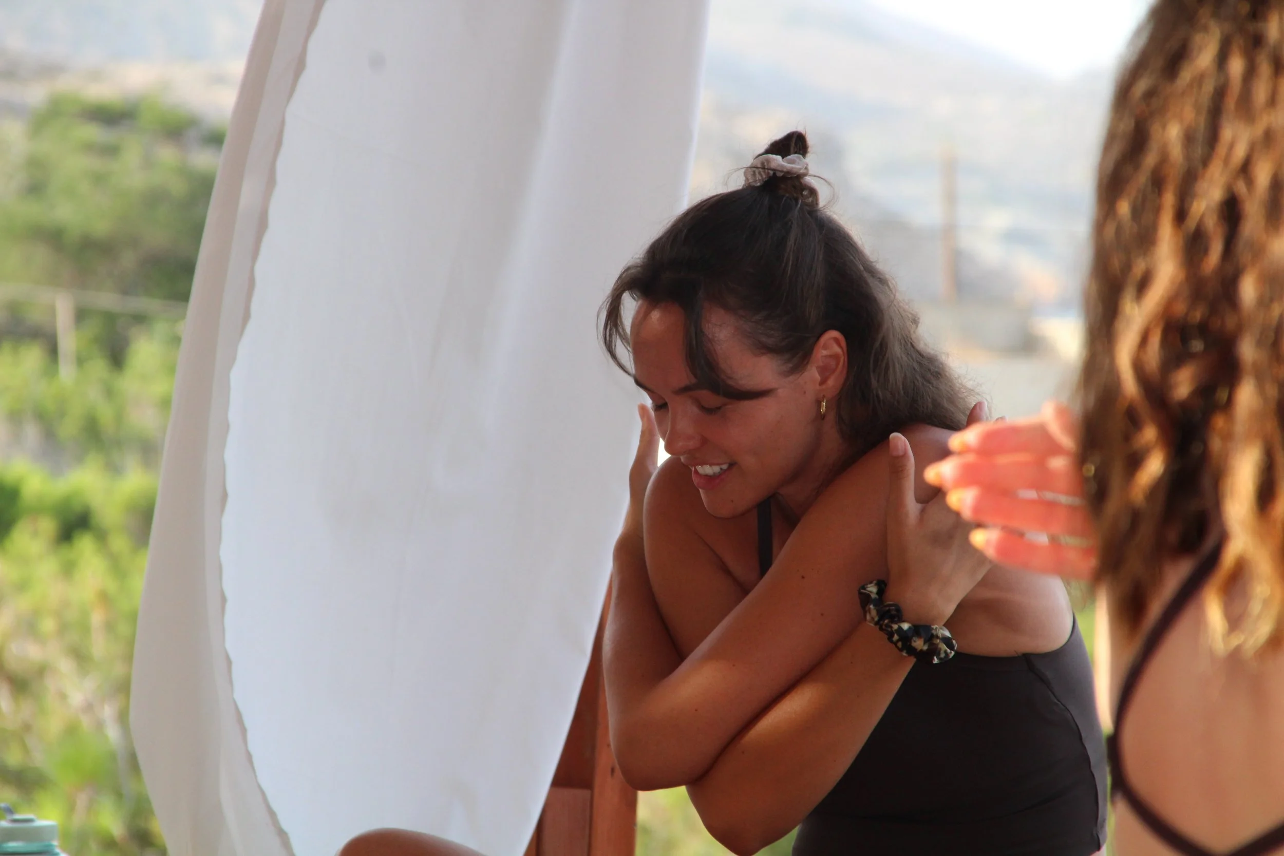 A woman with dark hair tied up, smiling and leaning her head on her arm, with her eyes closed, outdoors near a white structure, with another person partially visible on the right, and a background of greenery and blurred cityscape.