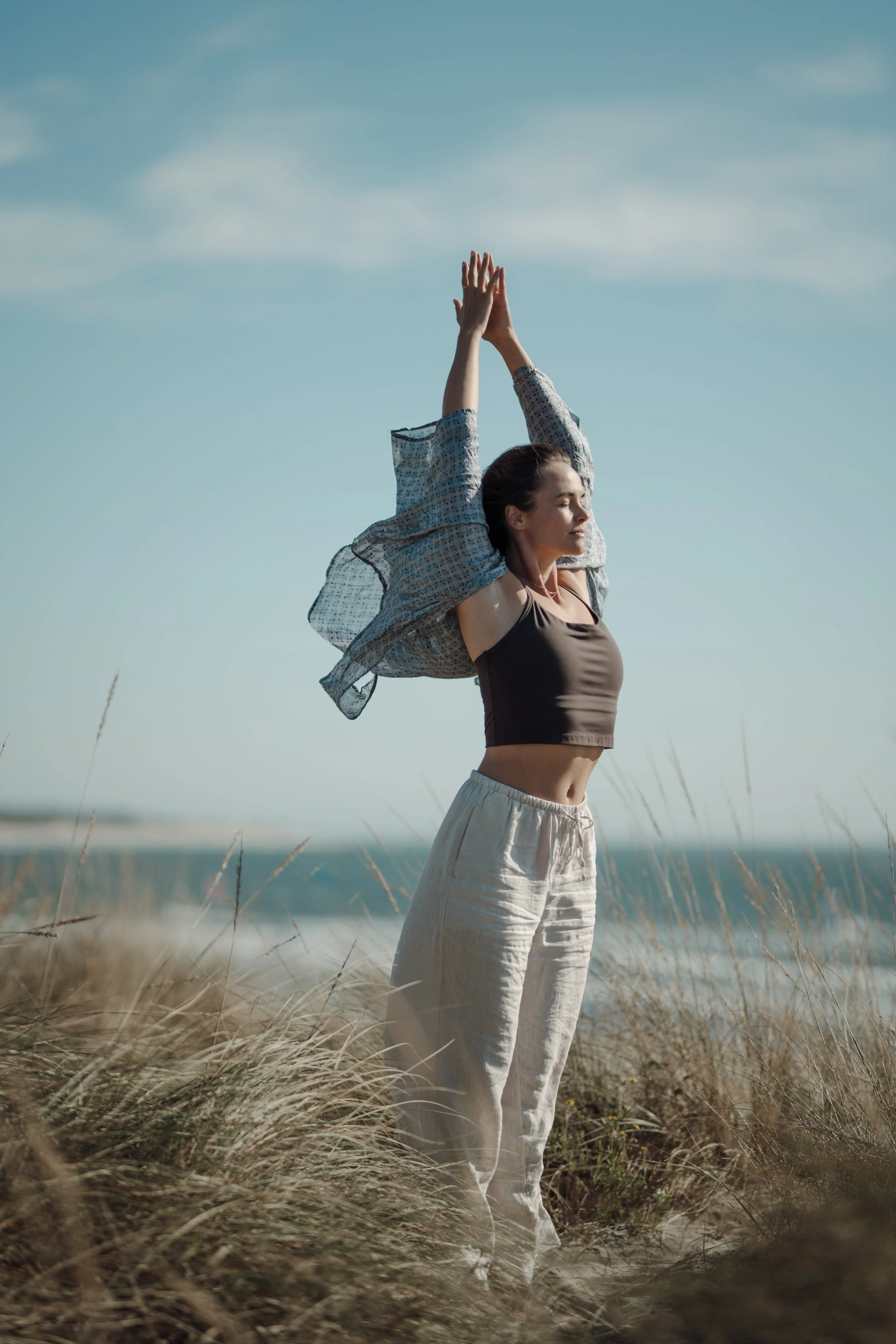 Woman standing with her arms raised in a yoga pose on a grassy hilltop near the beach with the ocean and sky in the background.