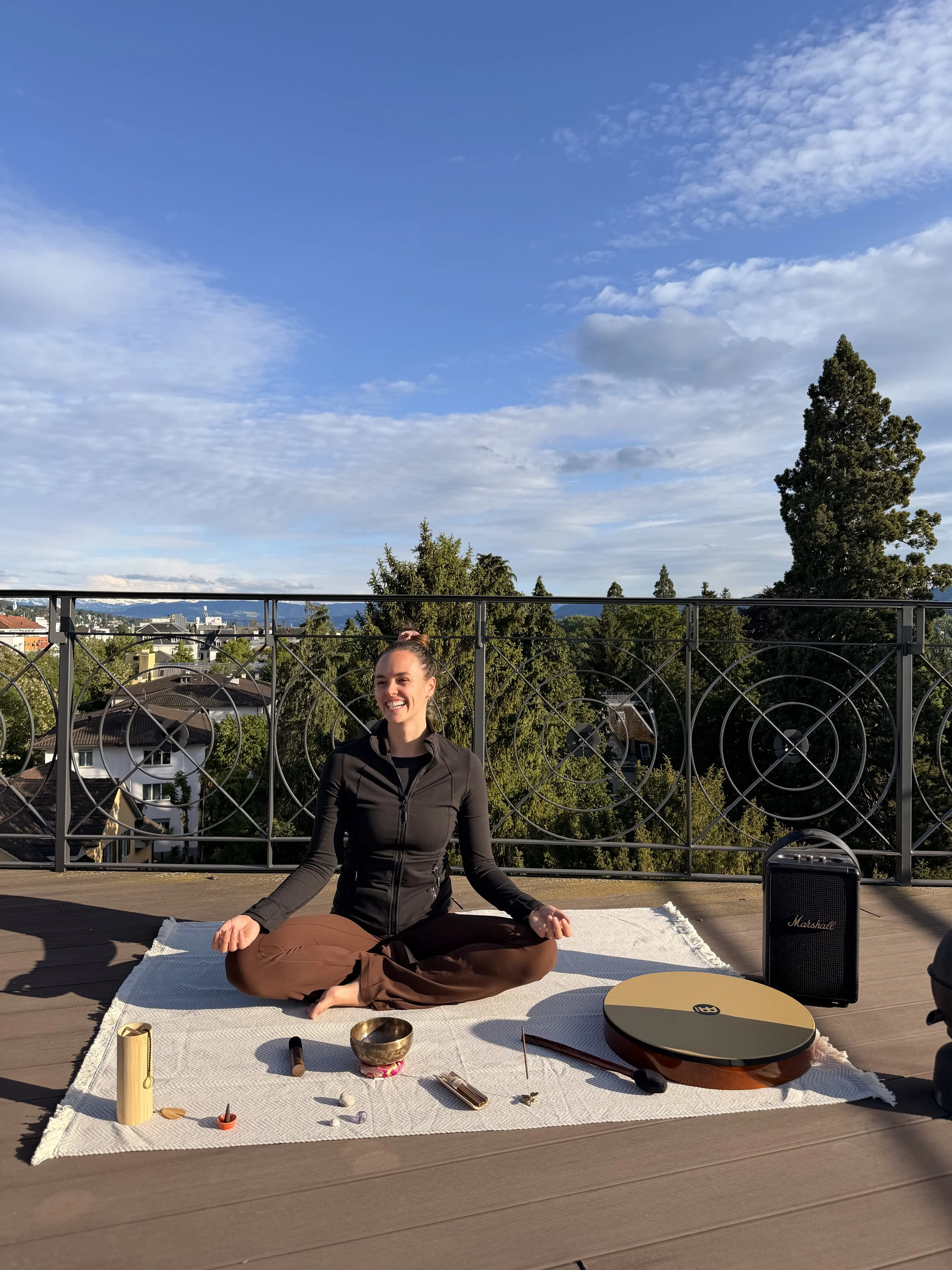 A woman practicing yoga outdoors on a rooftop with a view of trees, houses, and mountains under a partly cloudy sky, sitting cross-legged on a white mat with various musical instruments and objects in front of her.