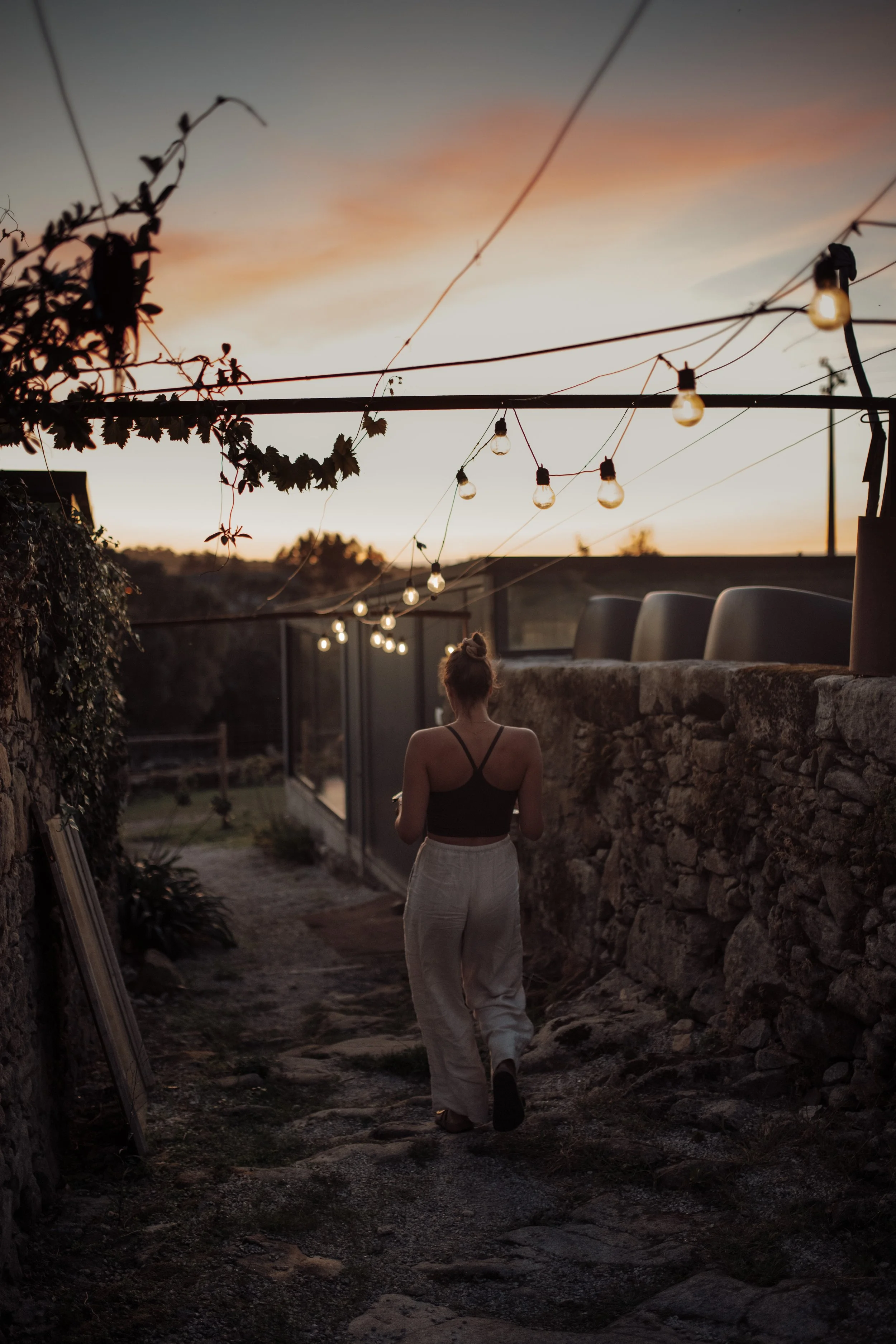 A woman walking along a stone path during sunset, with string lights hanging overhead and a stone wall on the right. The sky is painted with sunset colors, creating a warm, peaceful scene.