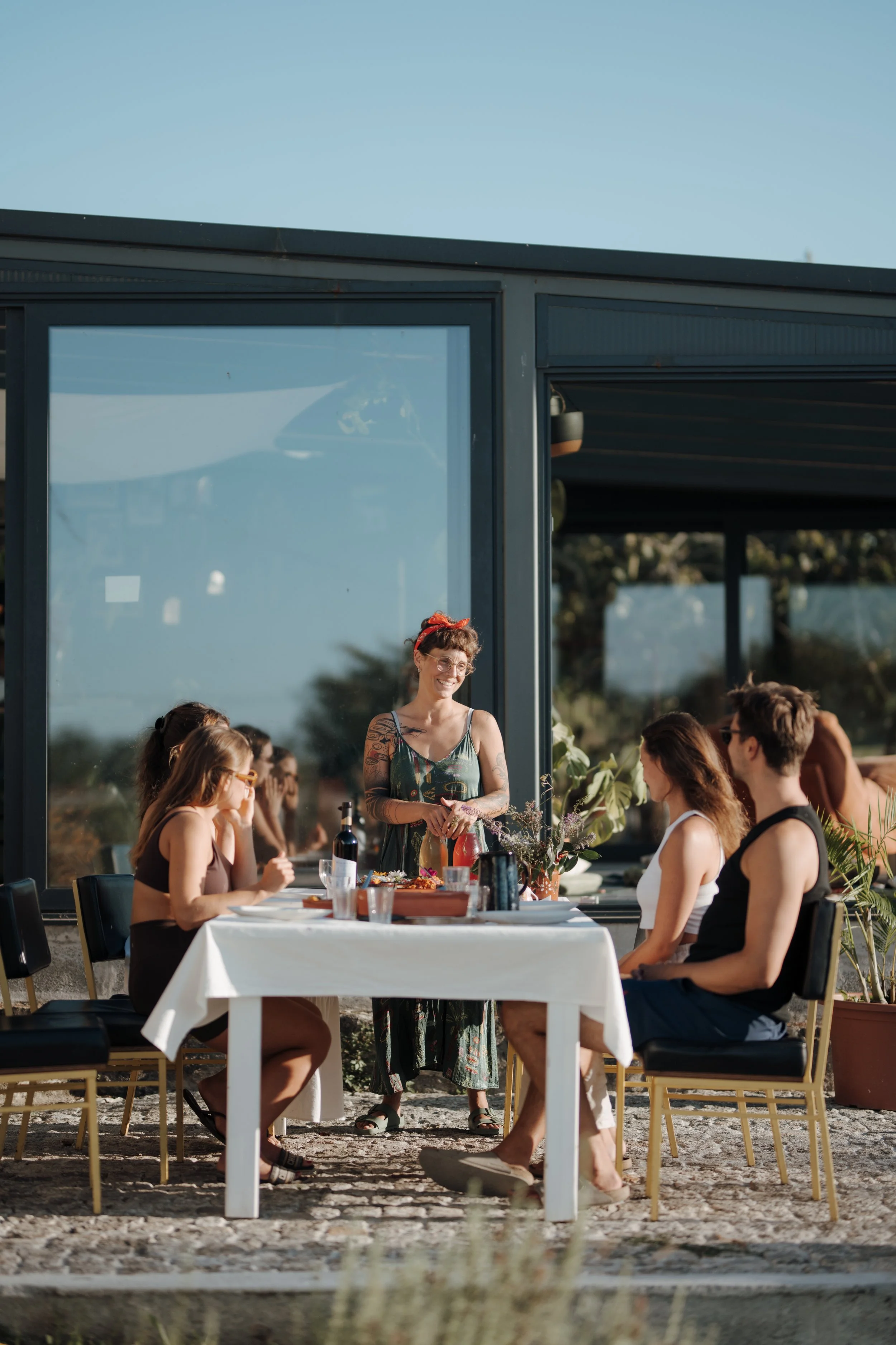 People gathered around a table outside, with a woman standing and speaking or presenting to the group. The scene is set against a modern building with large glass windows. The setting appears to be a social or outdoor dining event during the daytime.