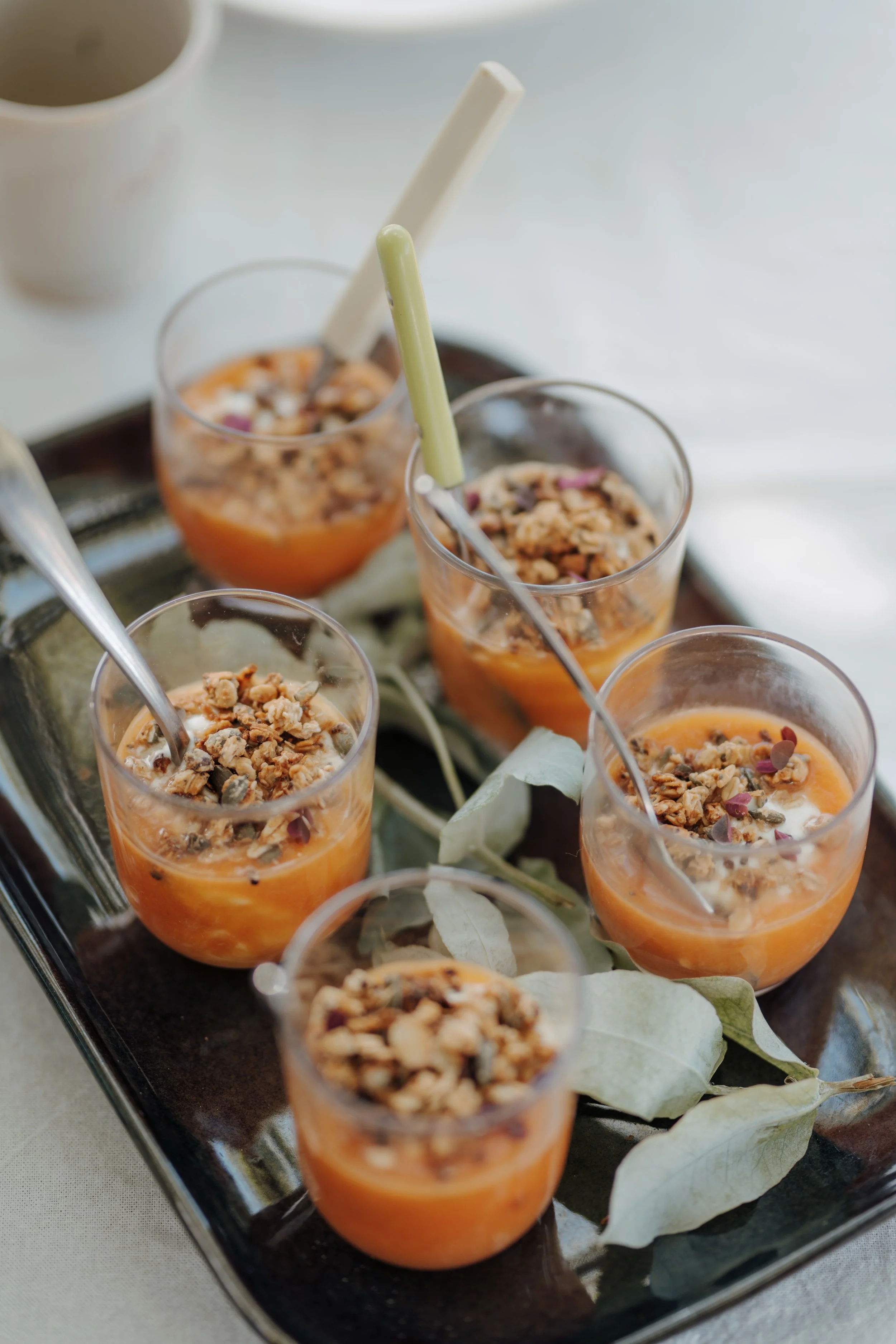Small glasses filled with an orange-colored dessert topped with granola and served on a black tray with decorative leaves.