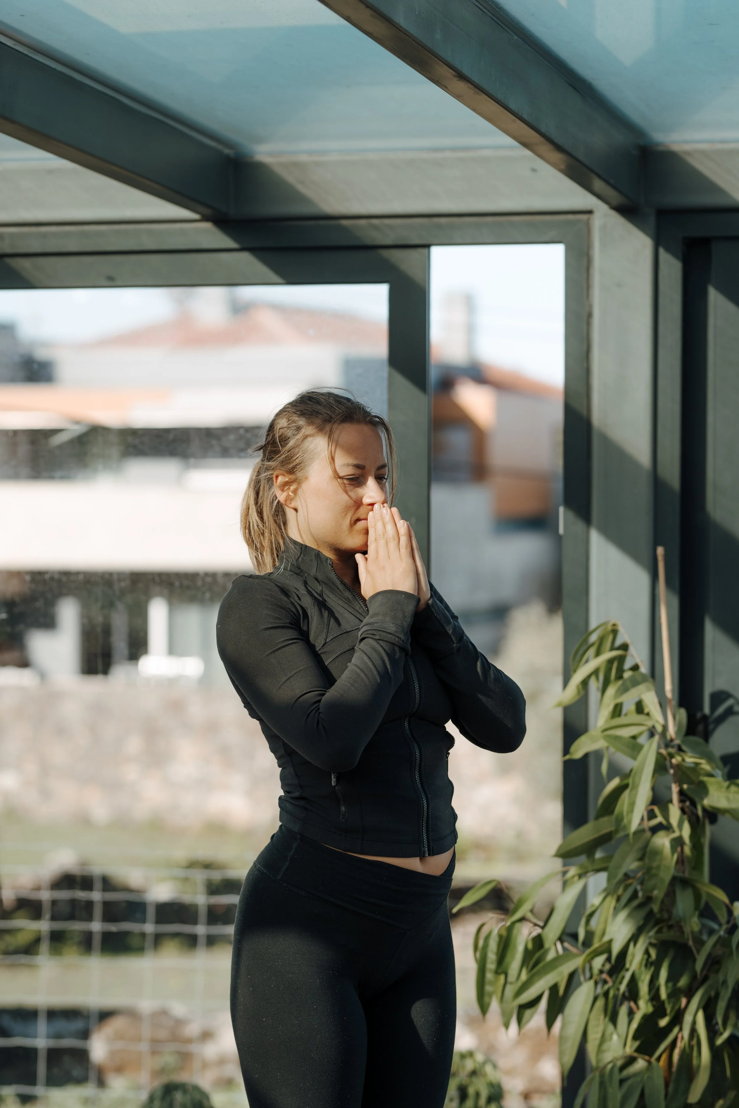 A woman in black athletic clothing practices yoga in a greenhouse, standing with her hands in prayer position near her face, with indoor plants nearby and a blurred outdoor background.