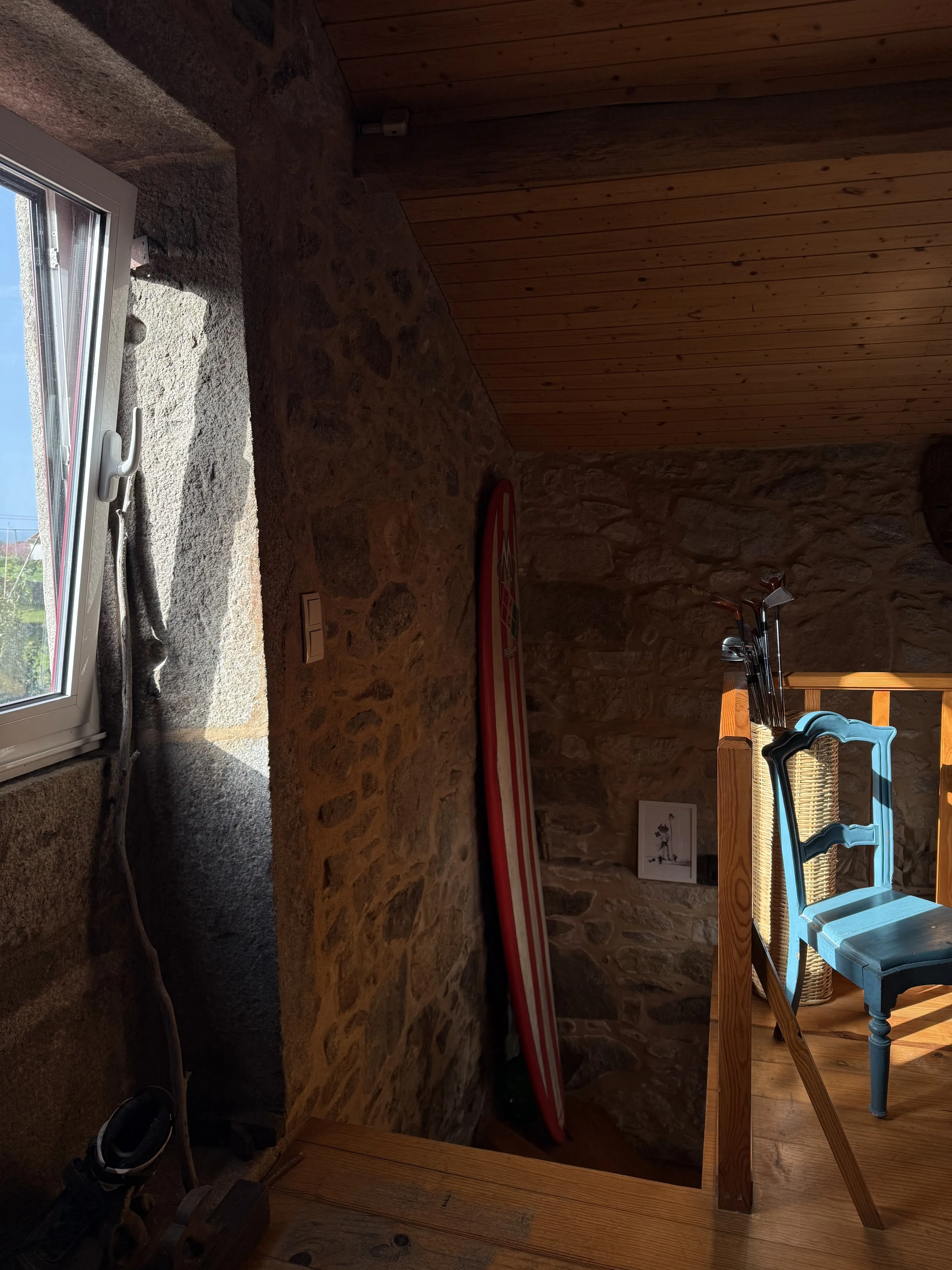 A room with stone wall and wooden ceiling. There are shoes on the floor near a window. A red and white surfboard leans against the stone wall. A wooden table with blue chairs and umbrellas is near the right side of the image.