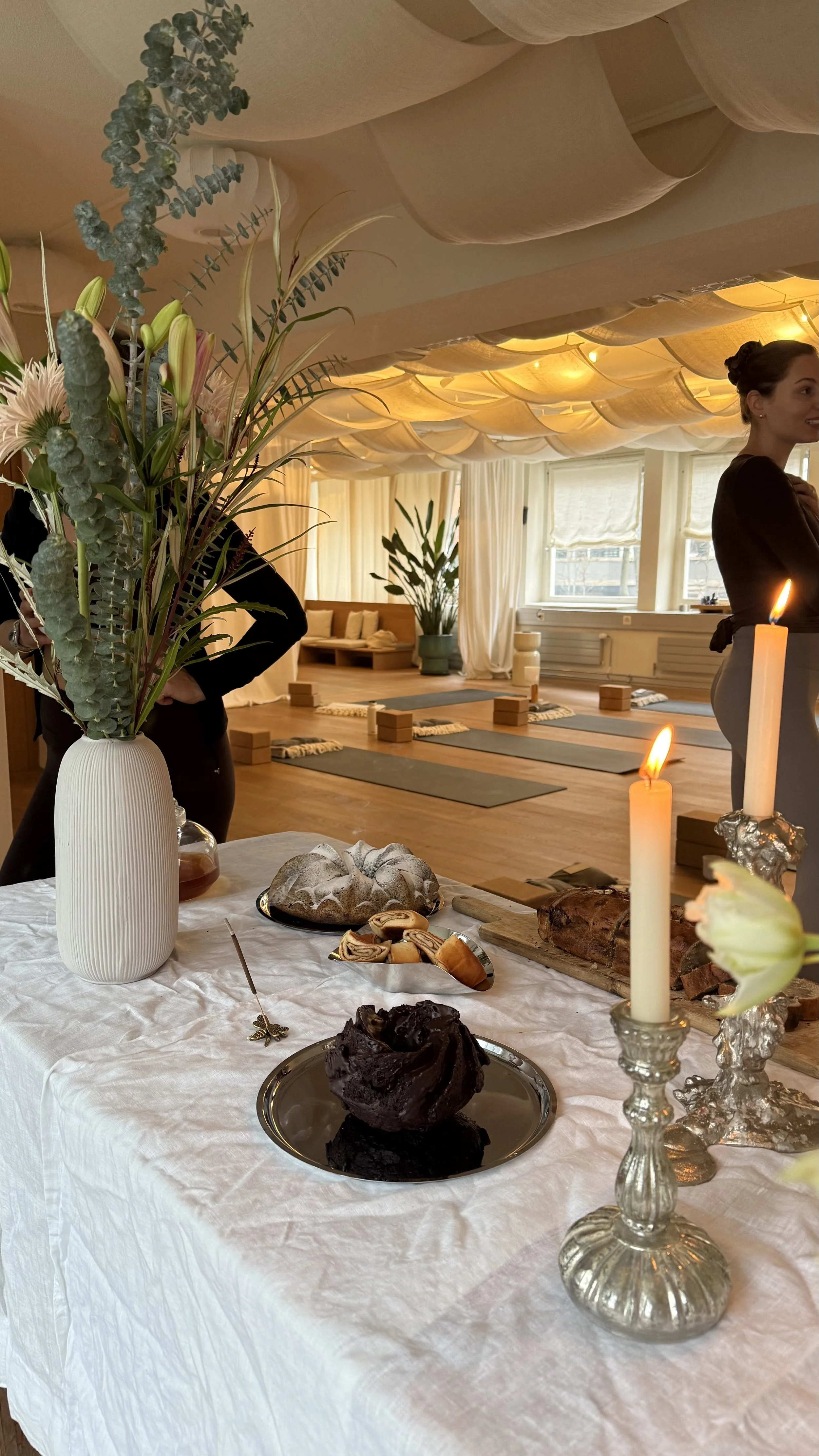 A table with a white tablecloth holding a vase with tall green and pink flowers, candles, pastries, and a chocolate cake. In the background, a yoga studio with mats, blocks, and a woman standing near windows with white curtains.