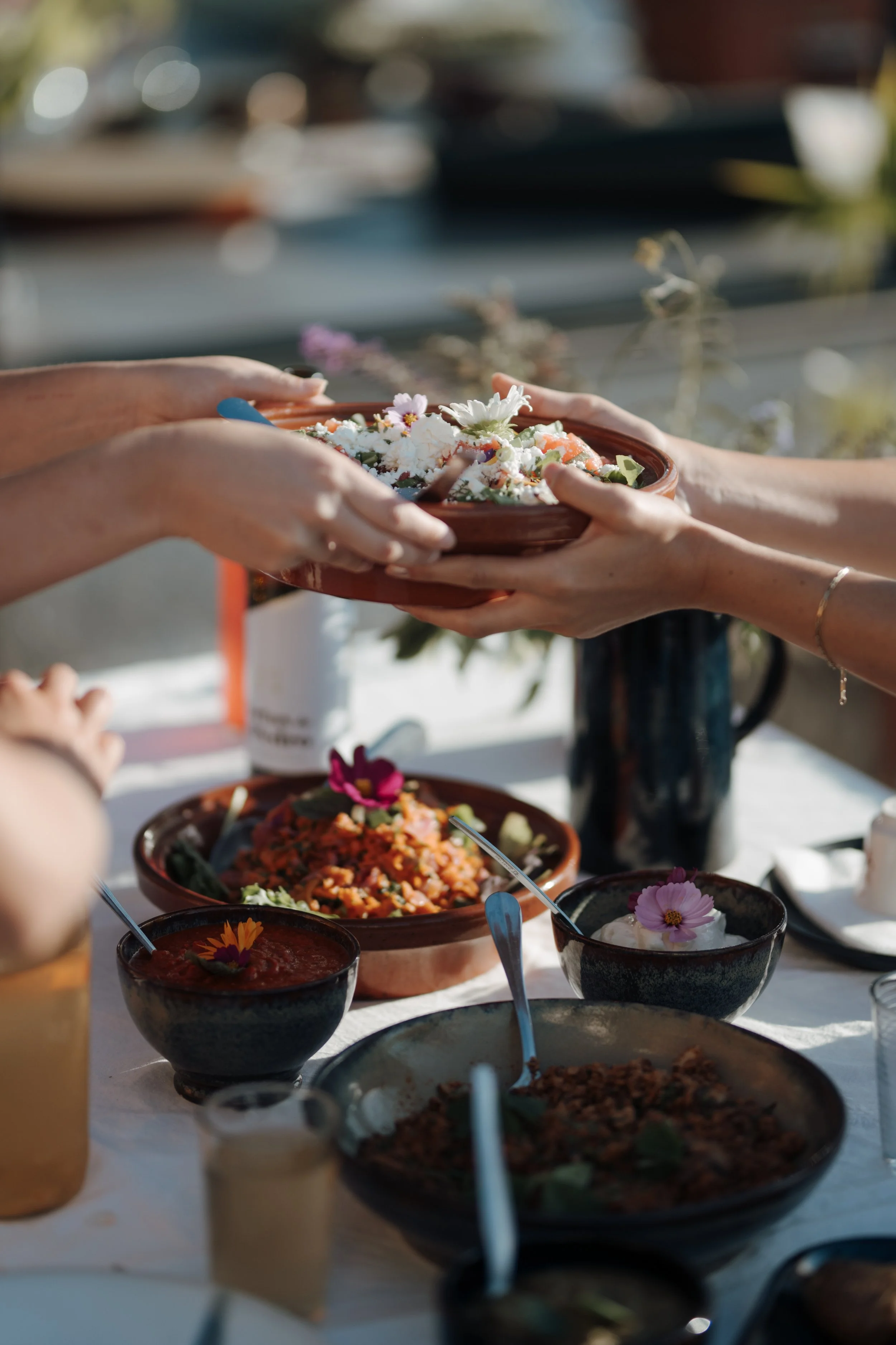 Hands of people exchanging a bowl of flowers and food at a table with various dishes and drinks during daylight.