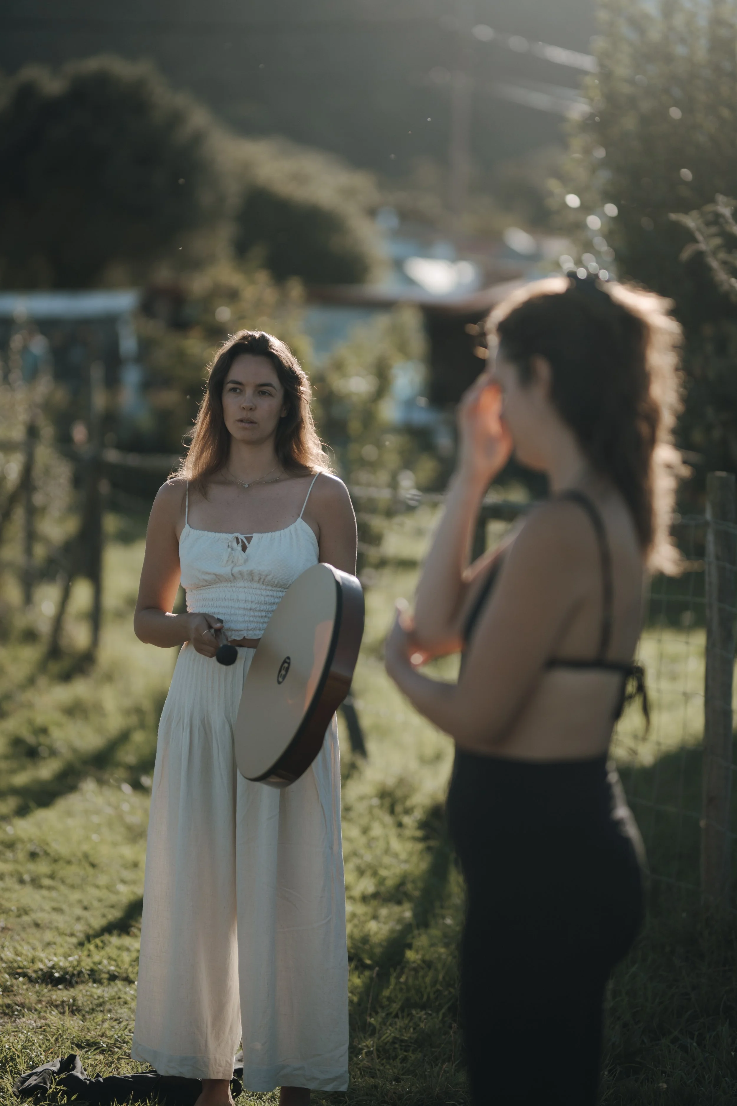 Two women standing outdoors on a grassy area, one holding a guitar body and the other touching her face, with trees and sunlight in the background.