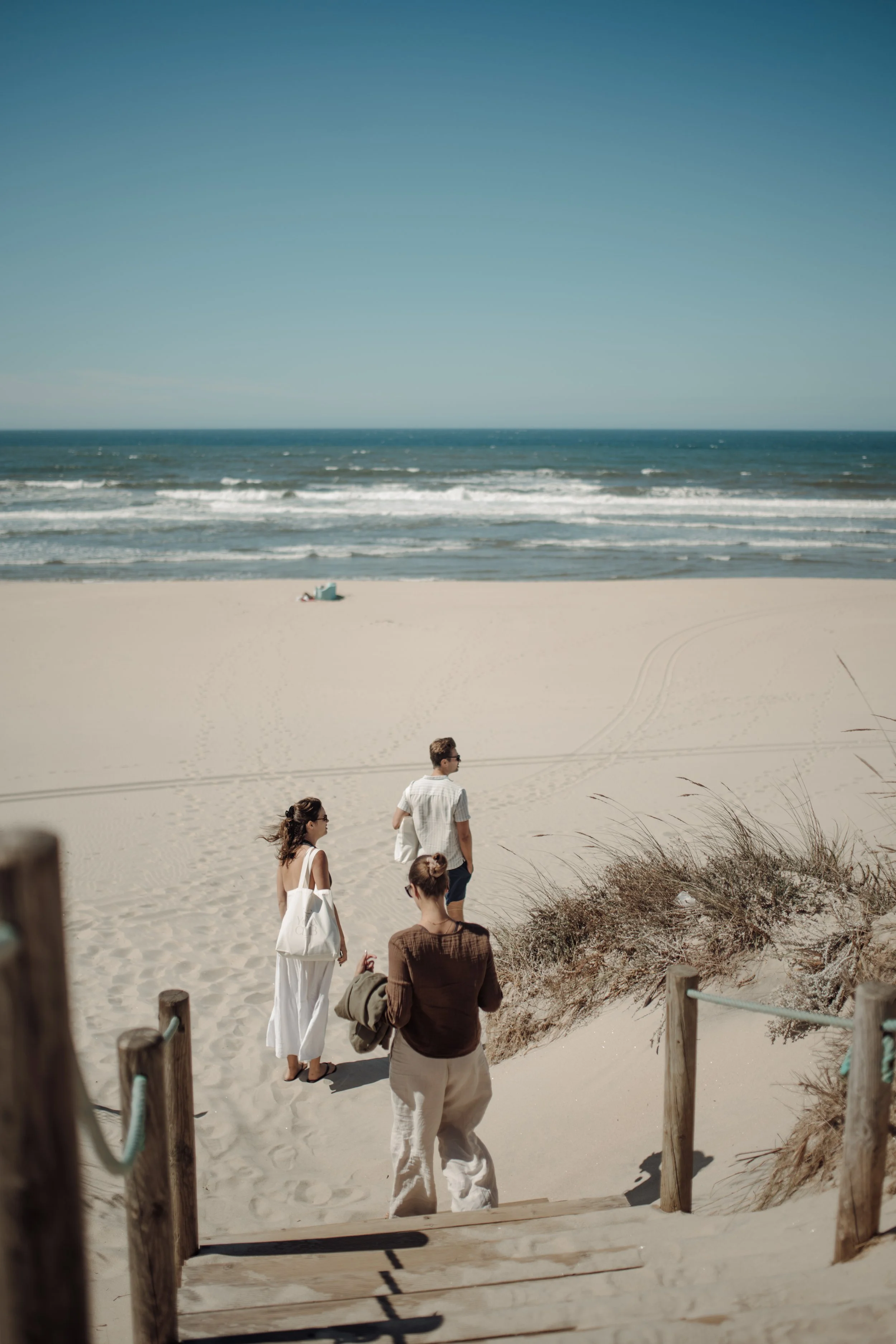 People walking down wooden steps onto a sandy beach with dunes, ocean waves, and a clear blue sky in the background.