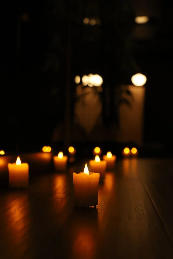 Several lit candles glowing softly on a dark surface, with more candles and blurry lighted objects in the background.