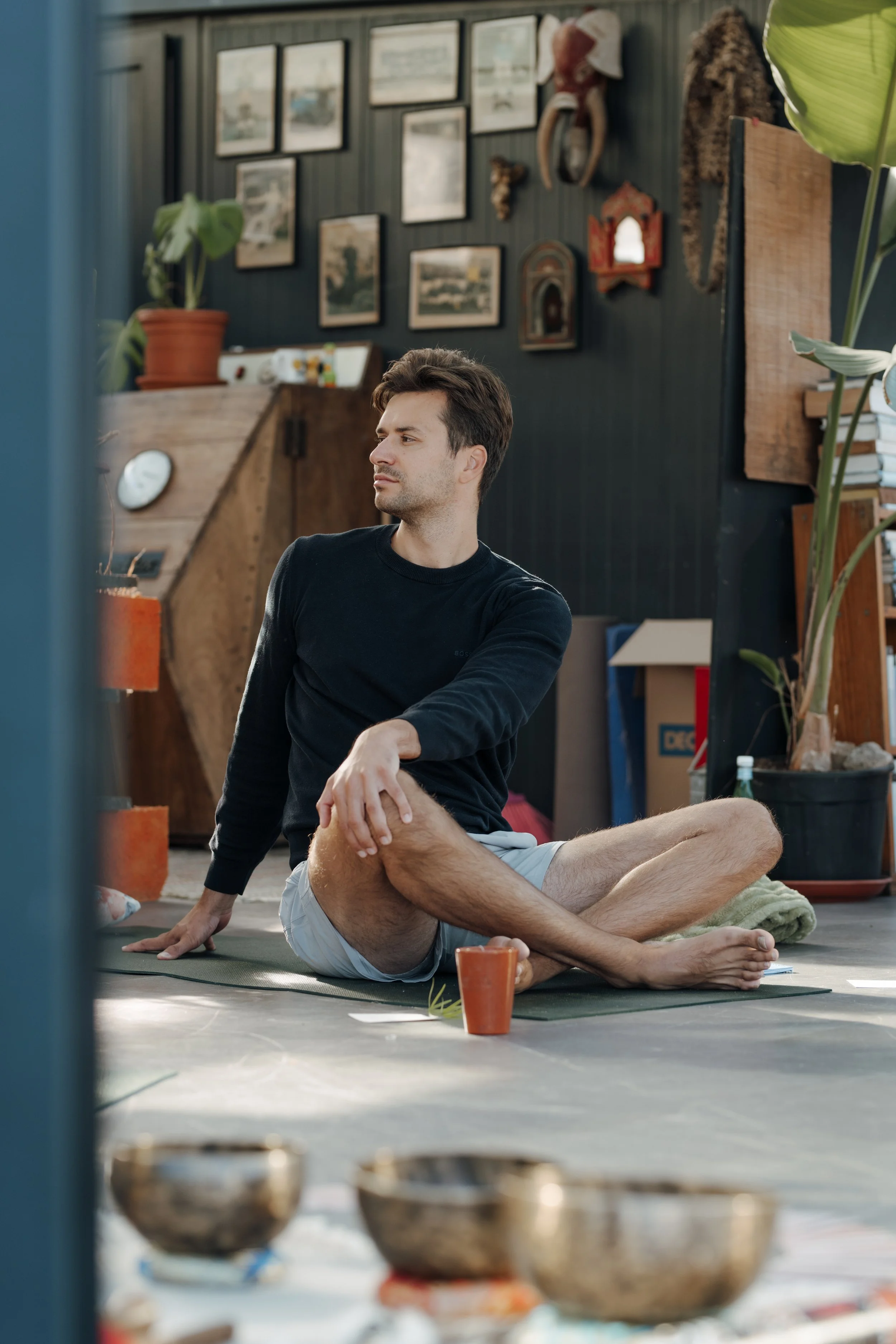 A man with brown hair and a serious expression, sitting cross-legged on a yoga mat indoors, with another person partially visible behind him, in a room decorated with pictures and artifacts on dark-colored walls, potted plants, and various objects.