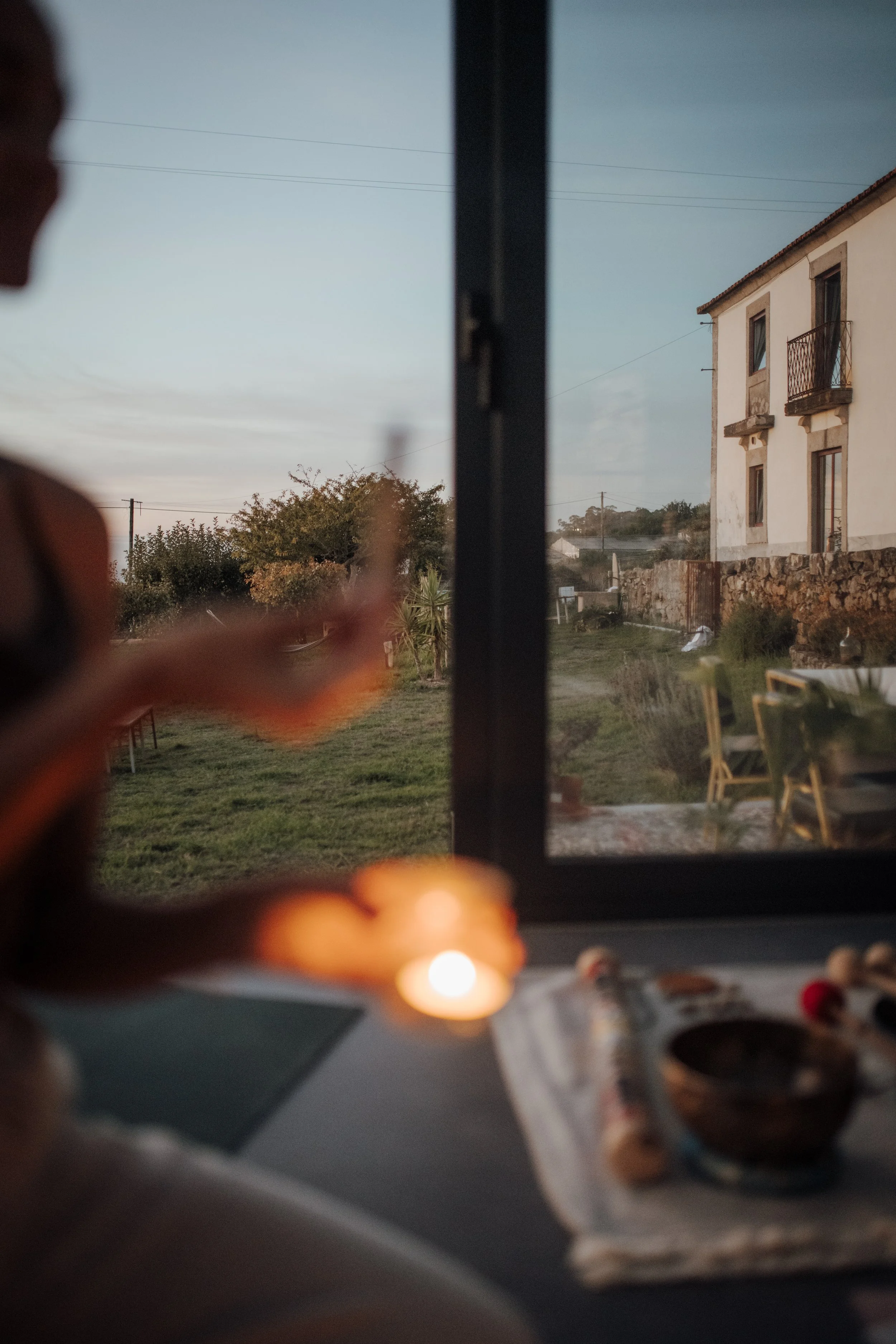 View through a window showing a house with a stone wall and a garden, with blurred objects and a candle in the foreground.
