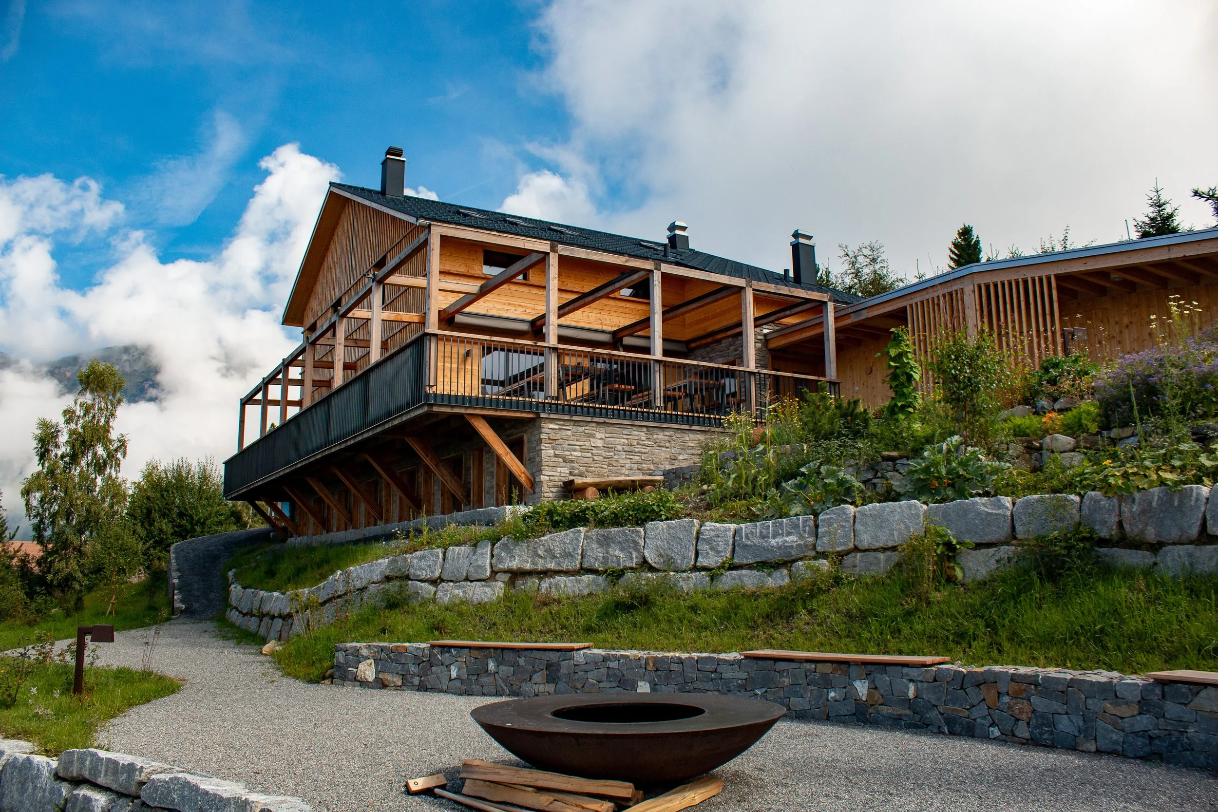 A modern wooden house on a hillside with a stone retaining wall, a deck, and a garden. The house has a black roof, multiple chimneys, and large windows. The sky is partly cloudy with blue sky visible.