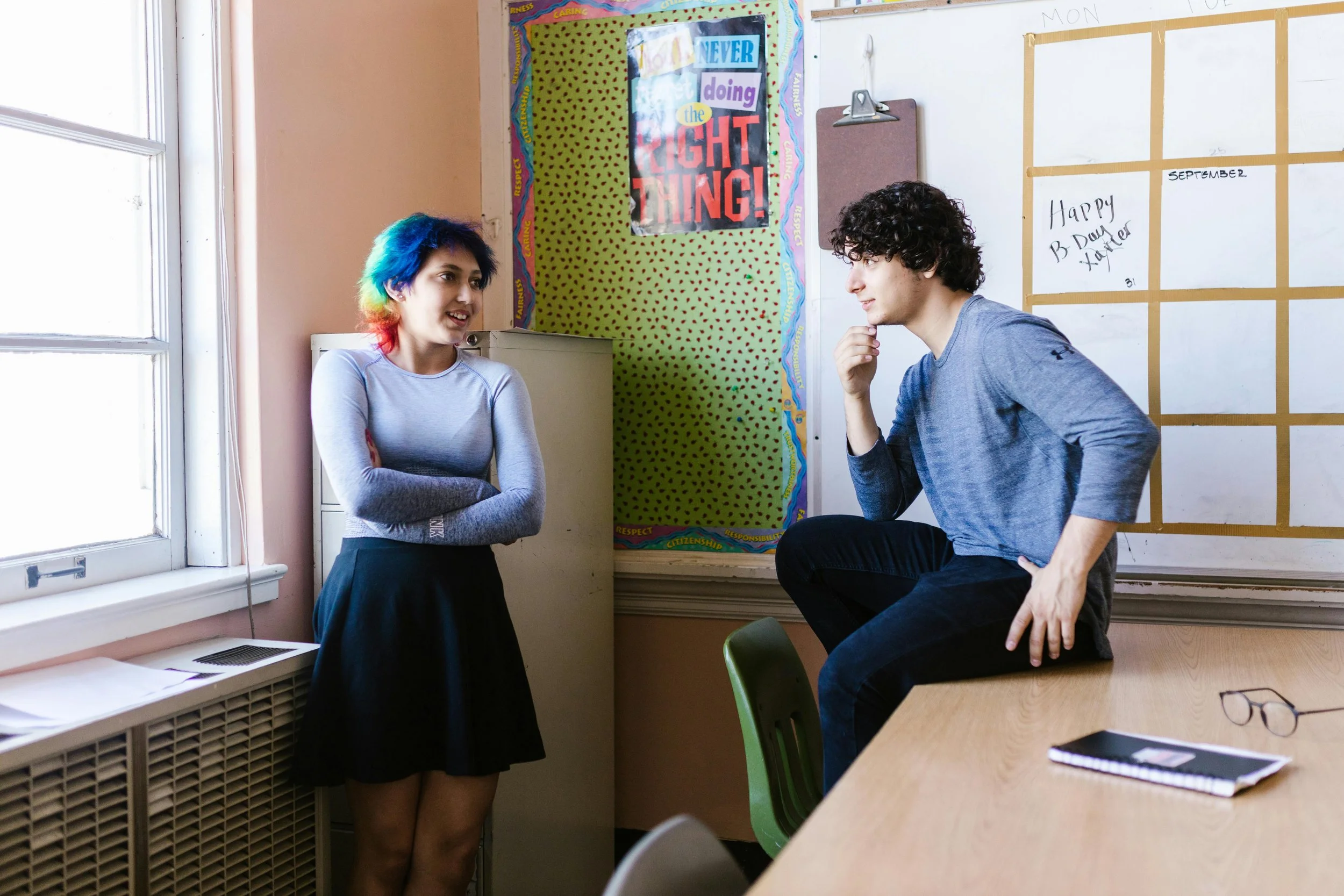 Classroom scene from Specialist High Skills Major Ontario: girl with rainbow hair near filing cabinet, boy pondering on desk by posters.