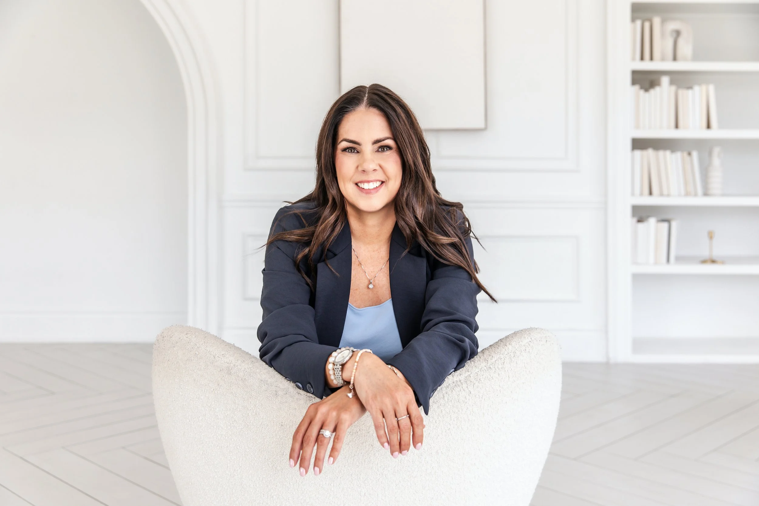 Danielle Pomeroy, a therapy provider in Ontario, smiling confidently in a navy blazer while leaning forward on a white chair in a modern white room.