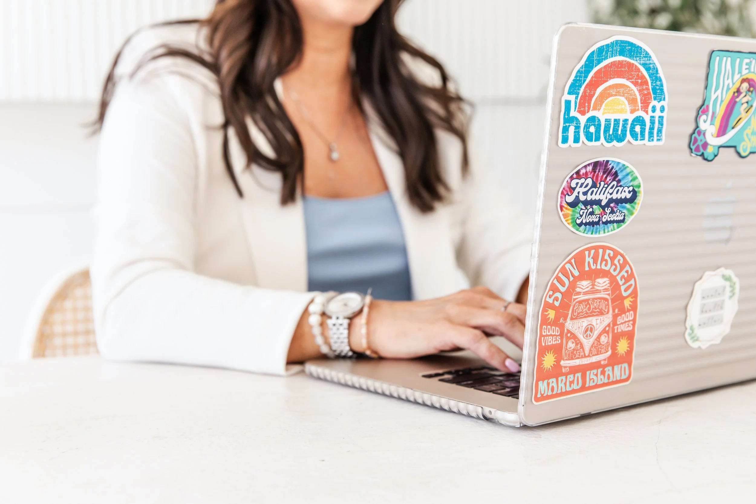 A Four Rooms Wellness mind body psychotherapist in a white blazer types on a sticker-covered laptop at a sunny desk.
