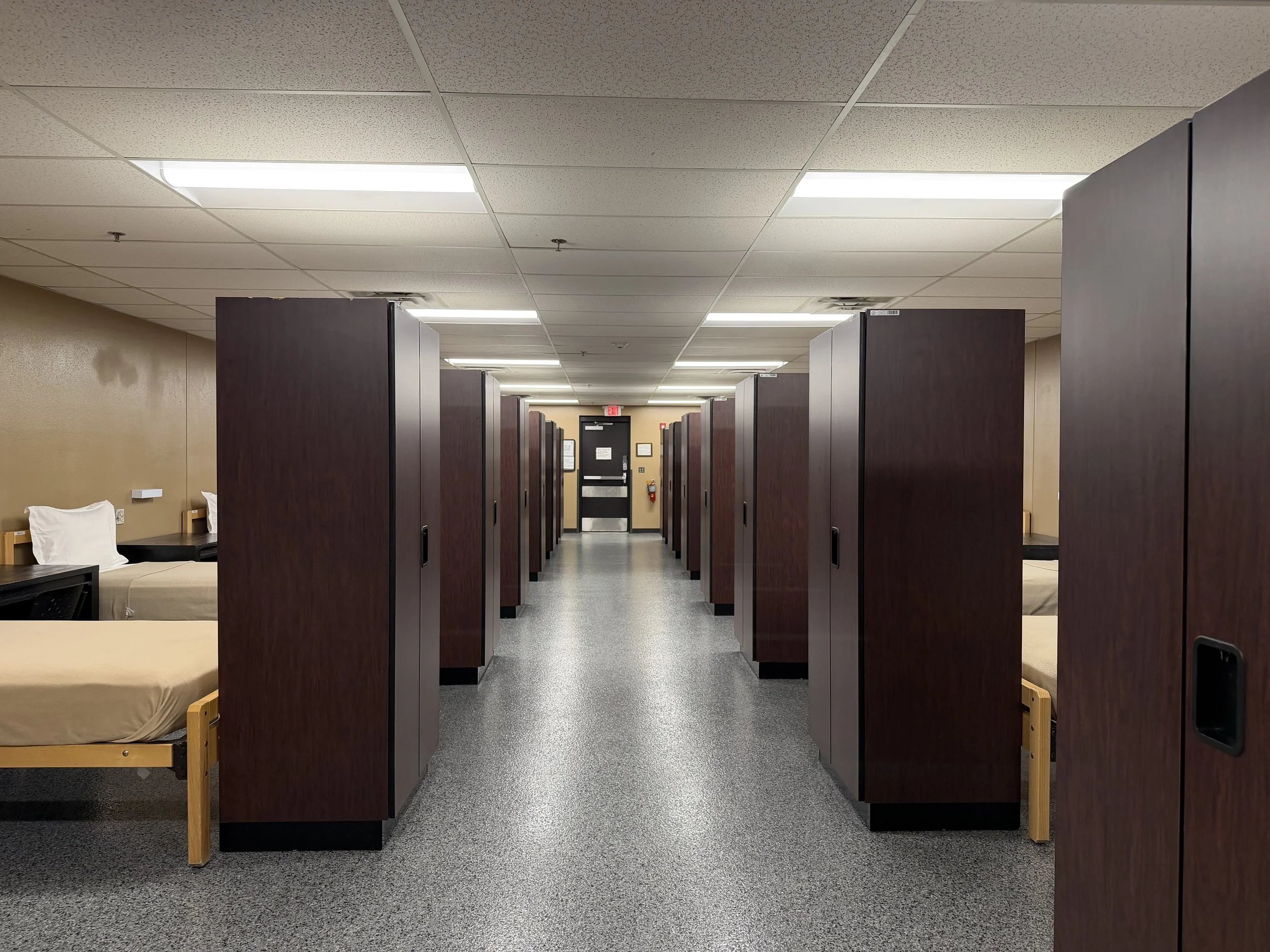 A walkway in an open bay with single beds separated by dark wood lockers.