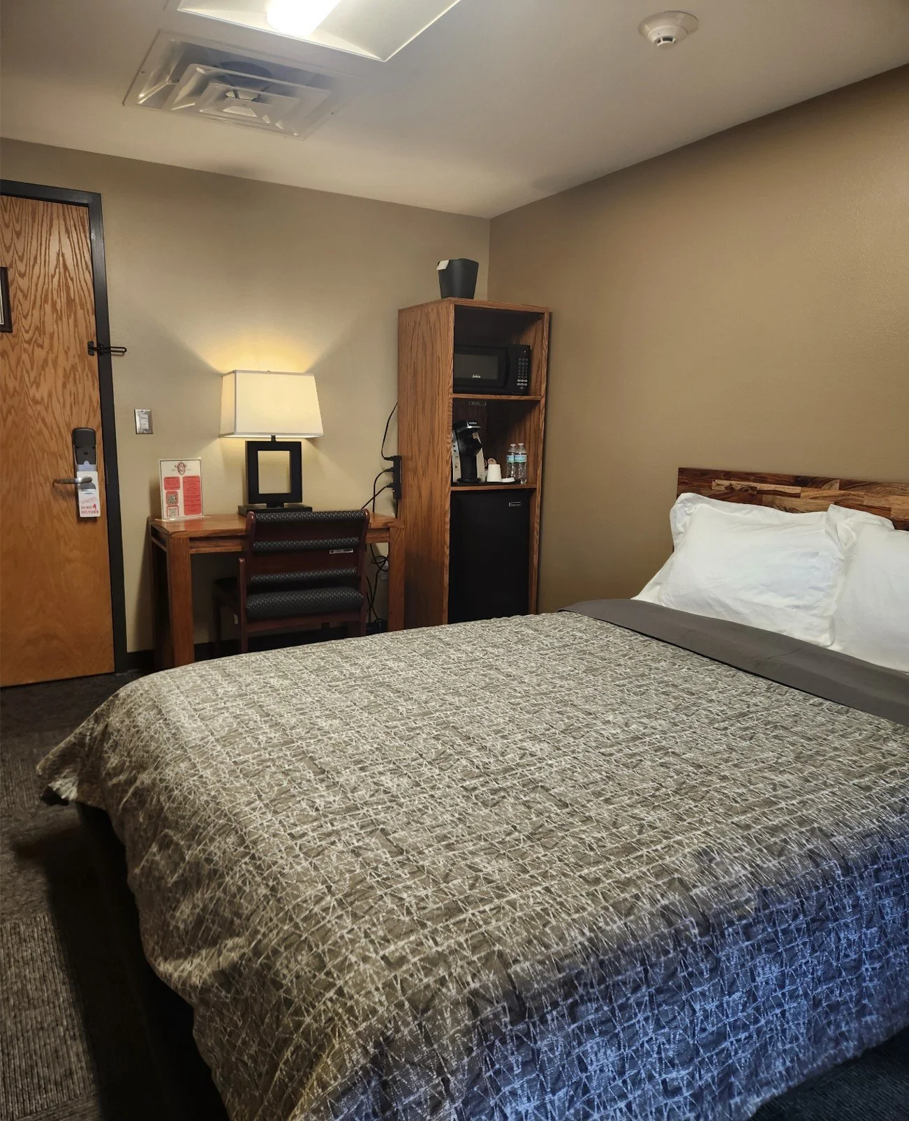 Interior of a hotel room with a bed, wooden headboard, pillows, a desk with a lamp, a chair, a tall wooden cabinet with a microwave, water bottles, a coffee maker, and a mini refrigerator, beige walls, and a smoke detector on the ceiling.
