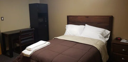 Hotel room with bed, pillows, bedside table, writing desk, chair, and mini fridge.