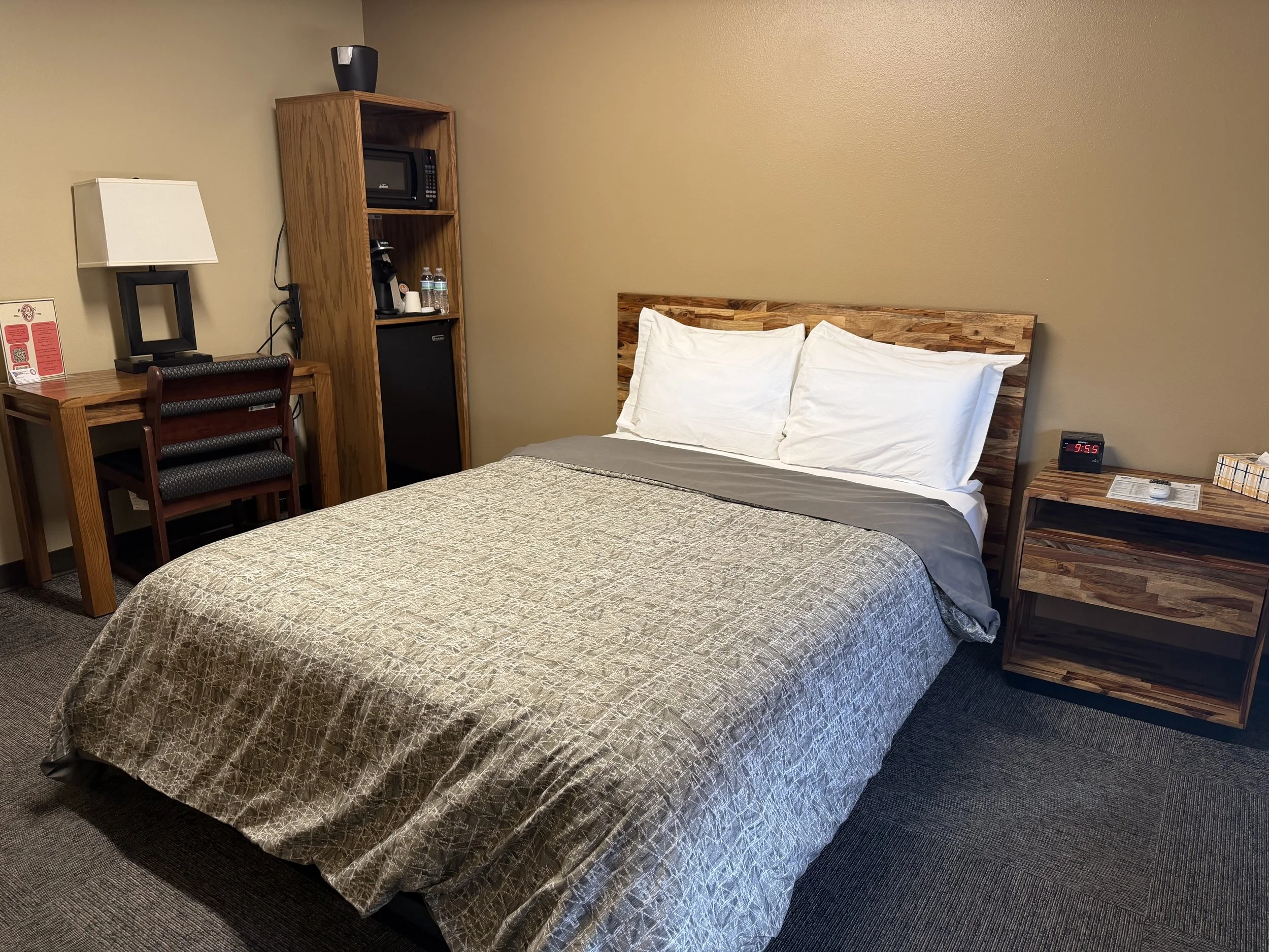 Hotel room with a bed, bedside table, desk with a lamp, chair, and a tall wooden shelf with a microwave, coffee maker, and water bottles.