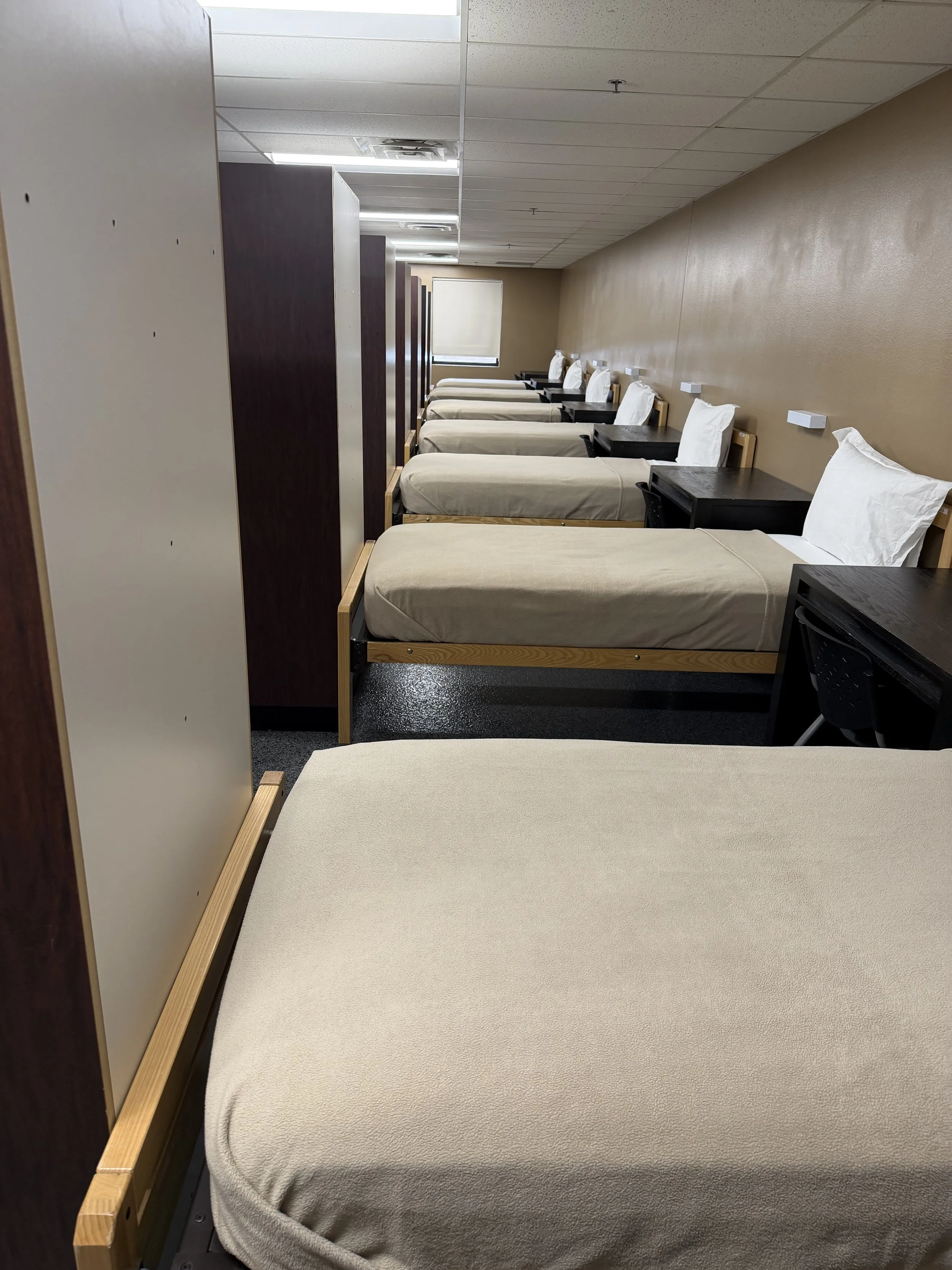 A row of hospital beds with beige mattresses, pillows, and black bedside tables, separated by privacy curtains in a medical facility