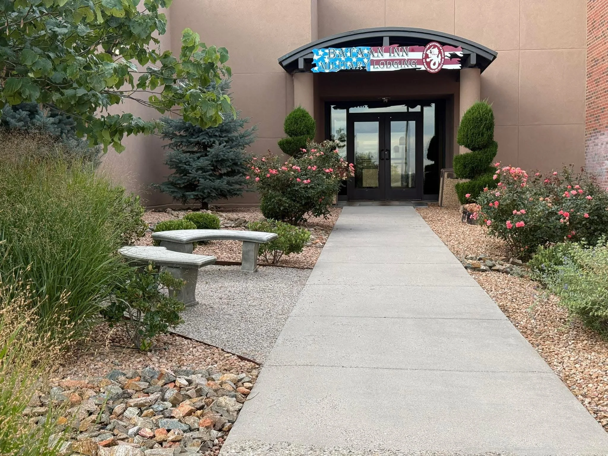 Entrance to the Bataan Inn Military Lodging, with a sidewalk leading to glass doors, surrounded by landscaped bushes and plants.