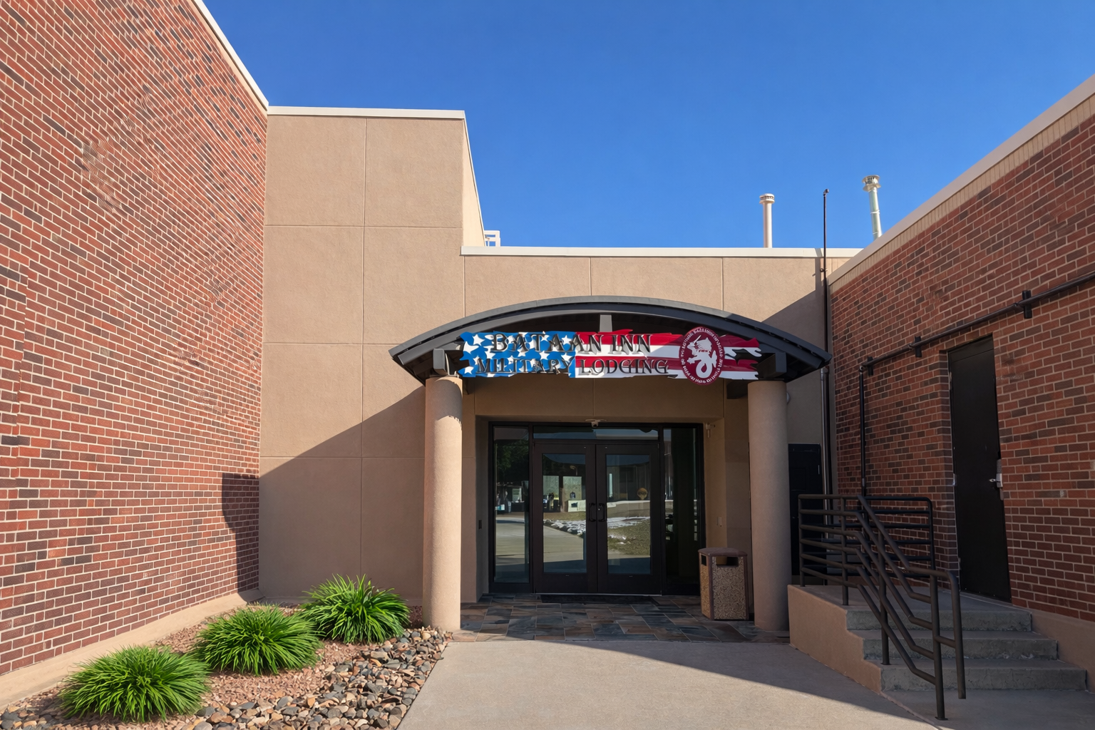 Entrance of a building with a sign reading 'Bataan Inn Military Lodging' with patriotic colors and symbols, glass doors, stairs, and landscaping with green bushes and rocks.