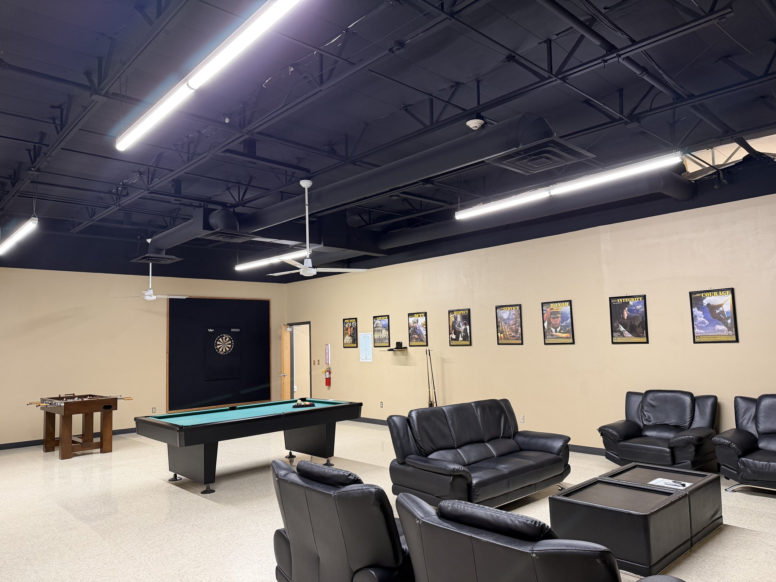 A game room with a pool table, dartboard, and sofa seating, decorated with motivational posters and kept illuminated by overhead lighting.