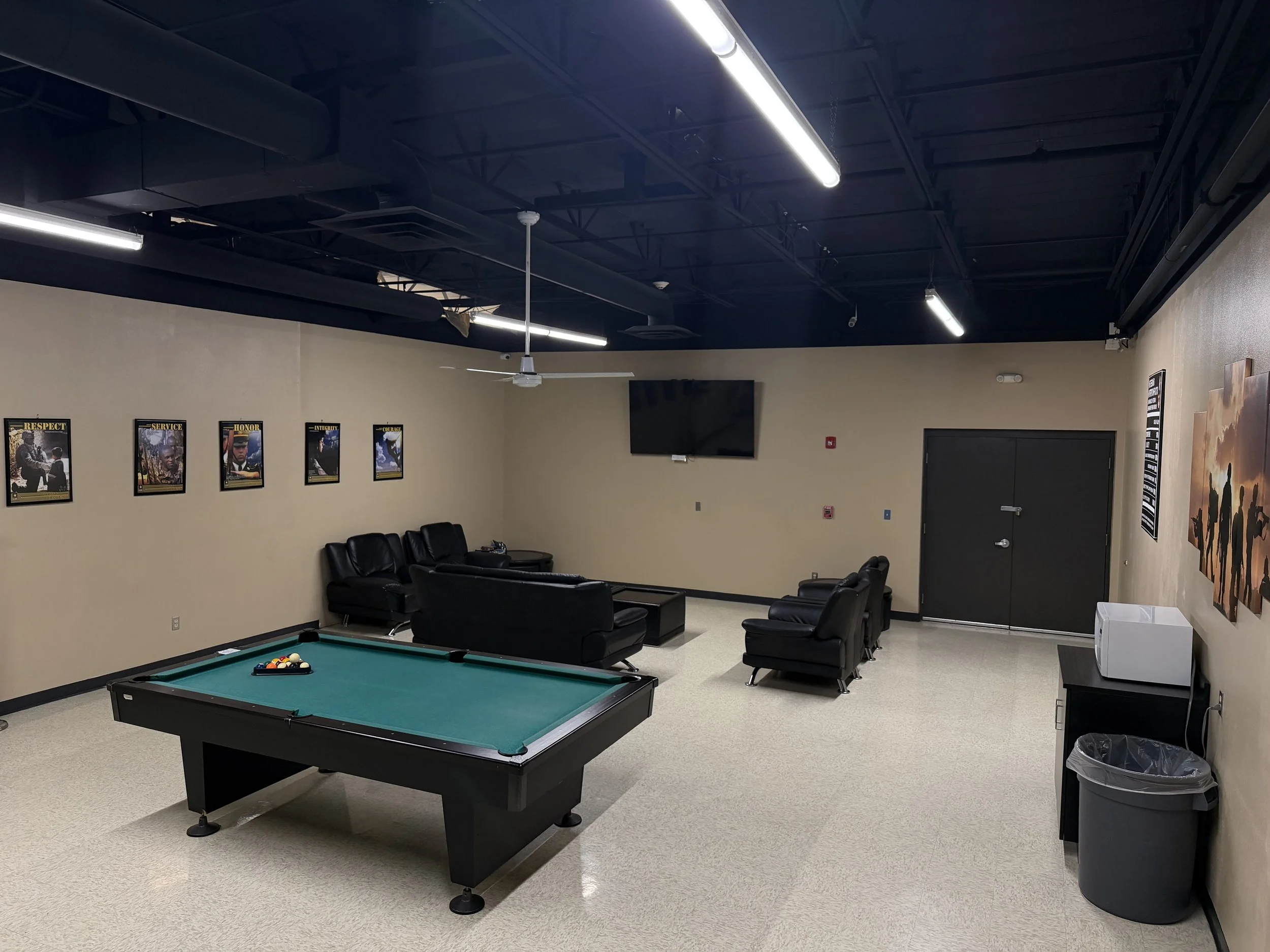 A lounge room with black ceiling and beige walls decorated with framed posters, black leather sofa set, a pool table, a flat-screen TV, and a small kitchenette with microwave and trash can.
